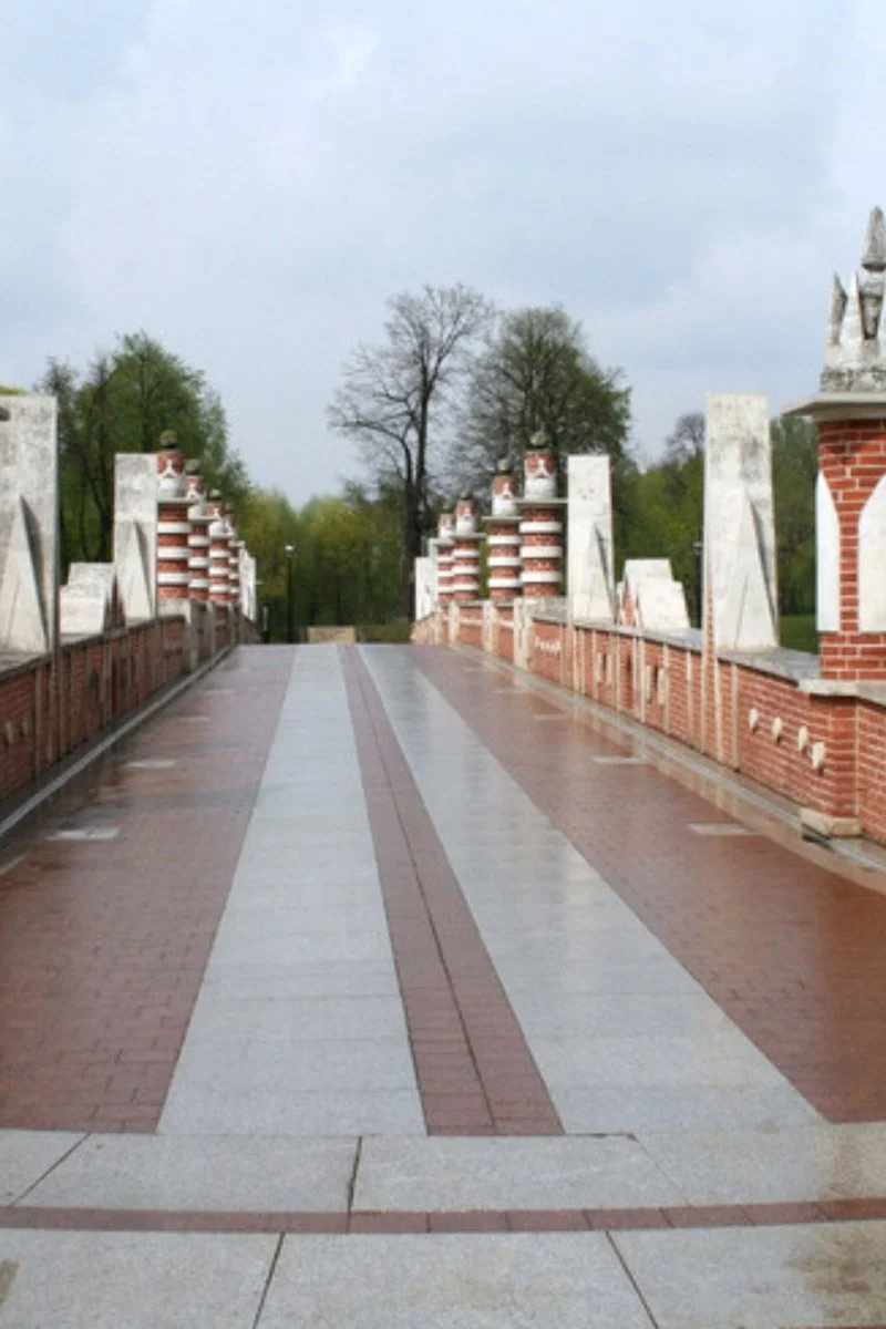 Empty brick and stone pedestrian bridge with decorative brick and stone archwork on the sides, in a park with trees in the background, overcast sky.