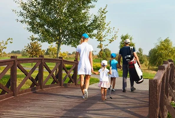 A woman with three children walking on a wooden bridge in a park or nature area, holding hands, with trees and clear sky in the background.
