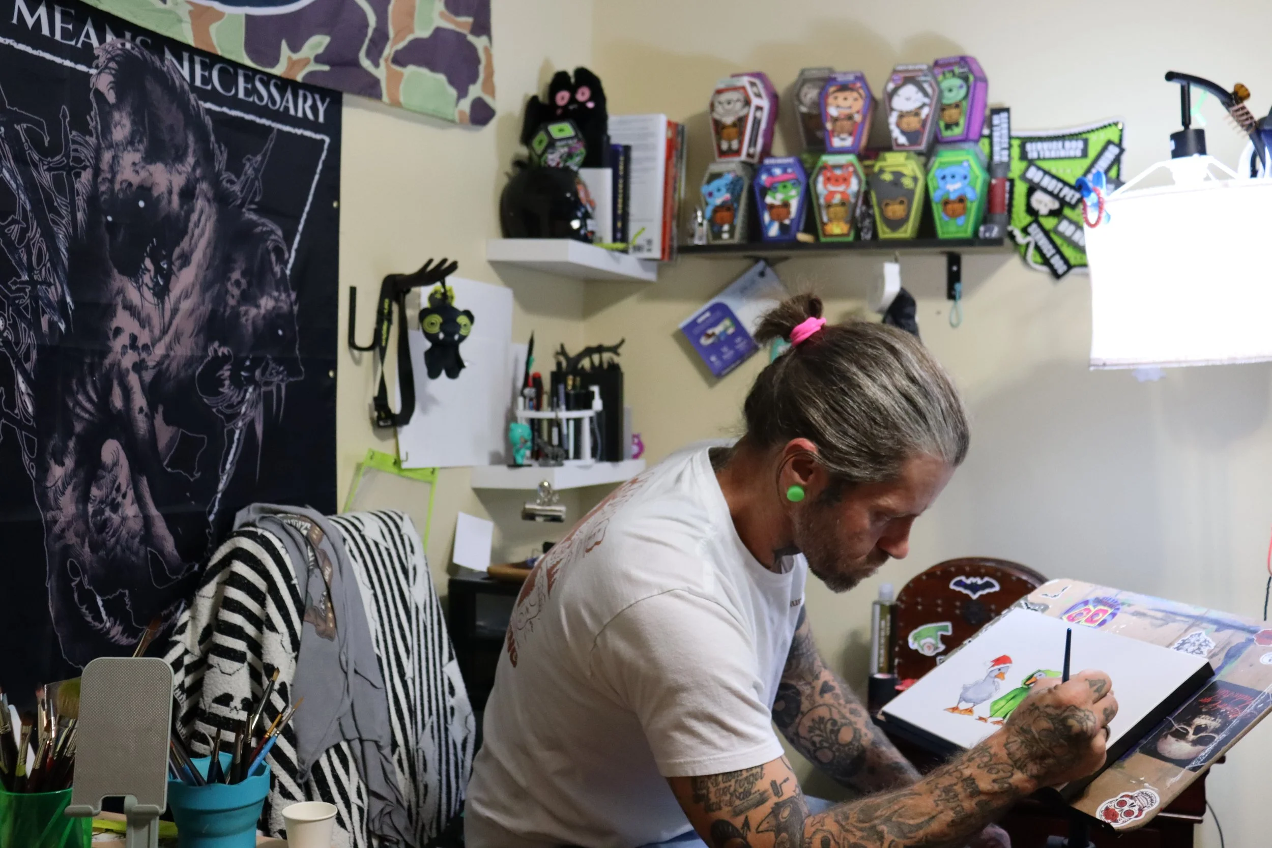 A man with tattoos and green earrings is drawing cartoon characters of a rooster and a duck on a sketchpad, seated at a cluttered desk in a room decorated with plush toys, posters, and art supplies.