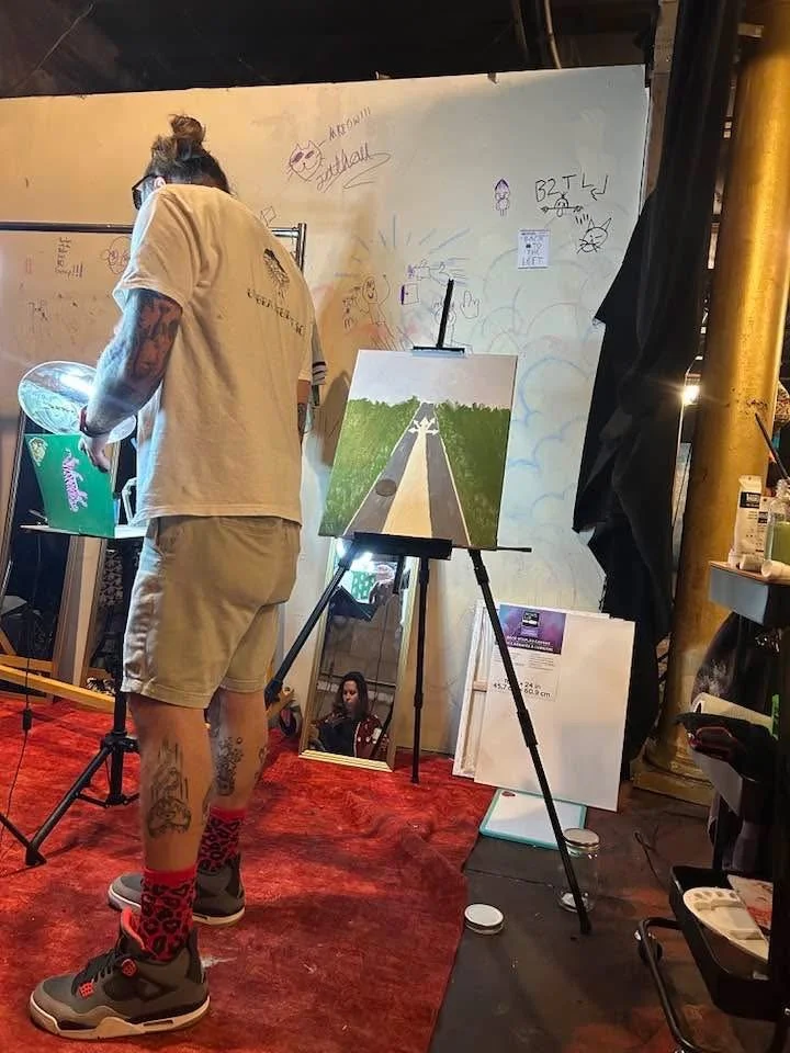An artist with tattoos on his arms and legs, wearing a cream t-shirt, beige shorts, gray sneakers, and red leopard print socks, is painting on a canvas that depicts a road leading into a forest during daytime. The studio has a red carpet and various framed pictures and artwork nearby, with some sketches and writings on a white wall in the background.