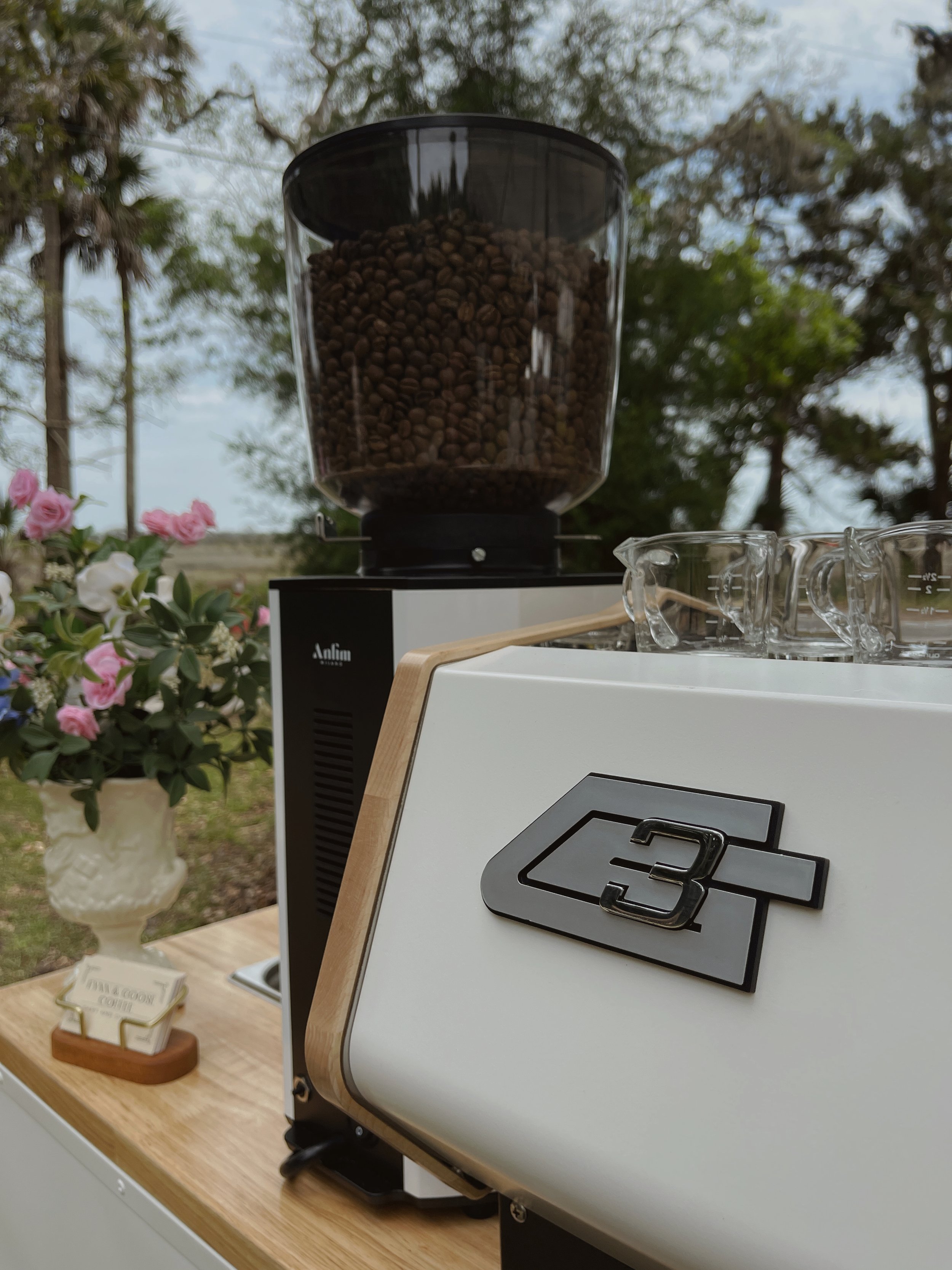 A coffee grinder filled with coffee beans, located outside on a wooden surface, with a white coffee machine in the foreground and pink and white flowers in a vase nearby.