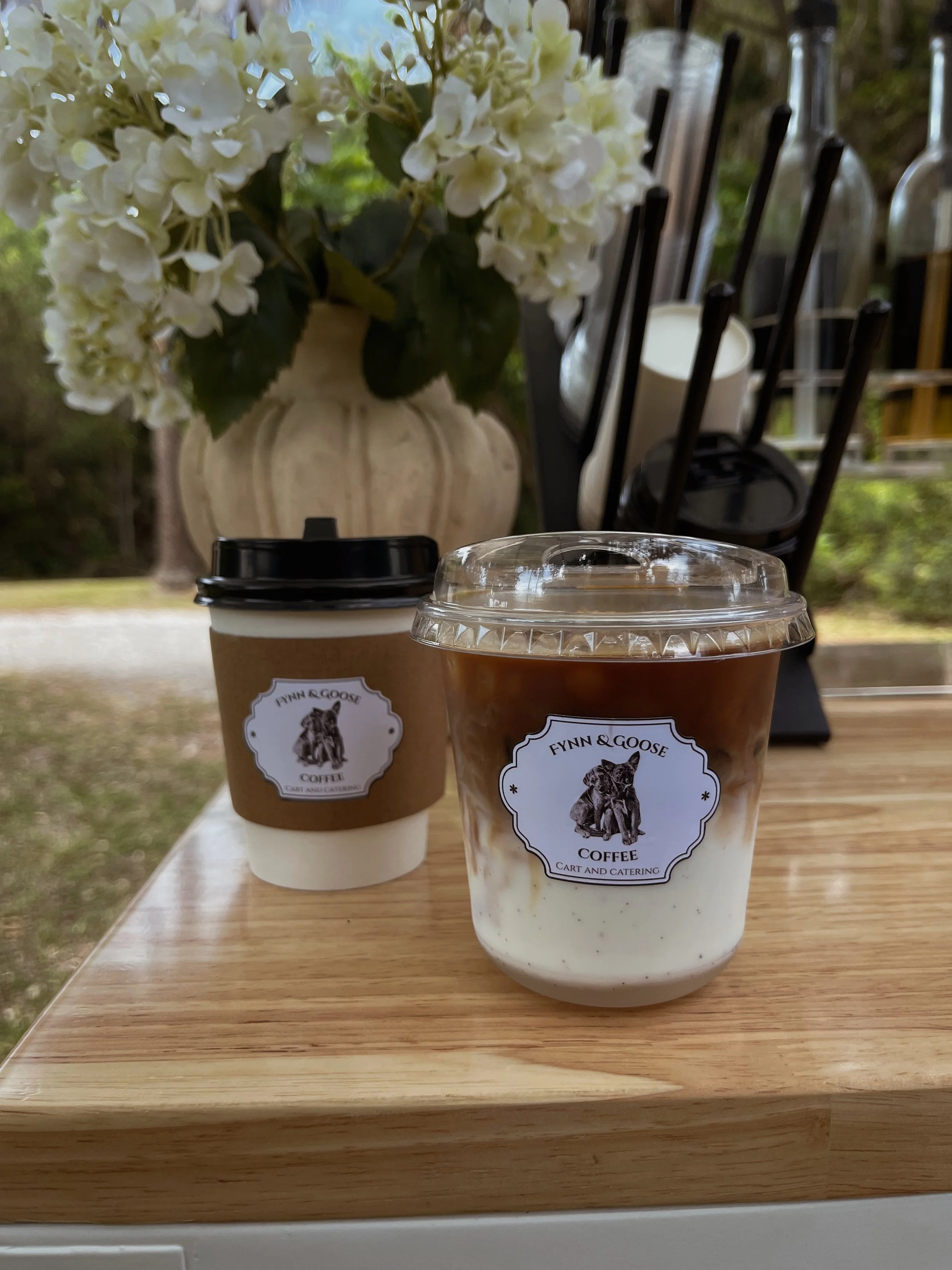 Two cups of coffee with foam and a logo labeled 'Fynn & GOOSE Coffee, Cart and Catering' on a wooden table, with a large floral arrangement of white hydrangeas in a beige vase in the background.