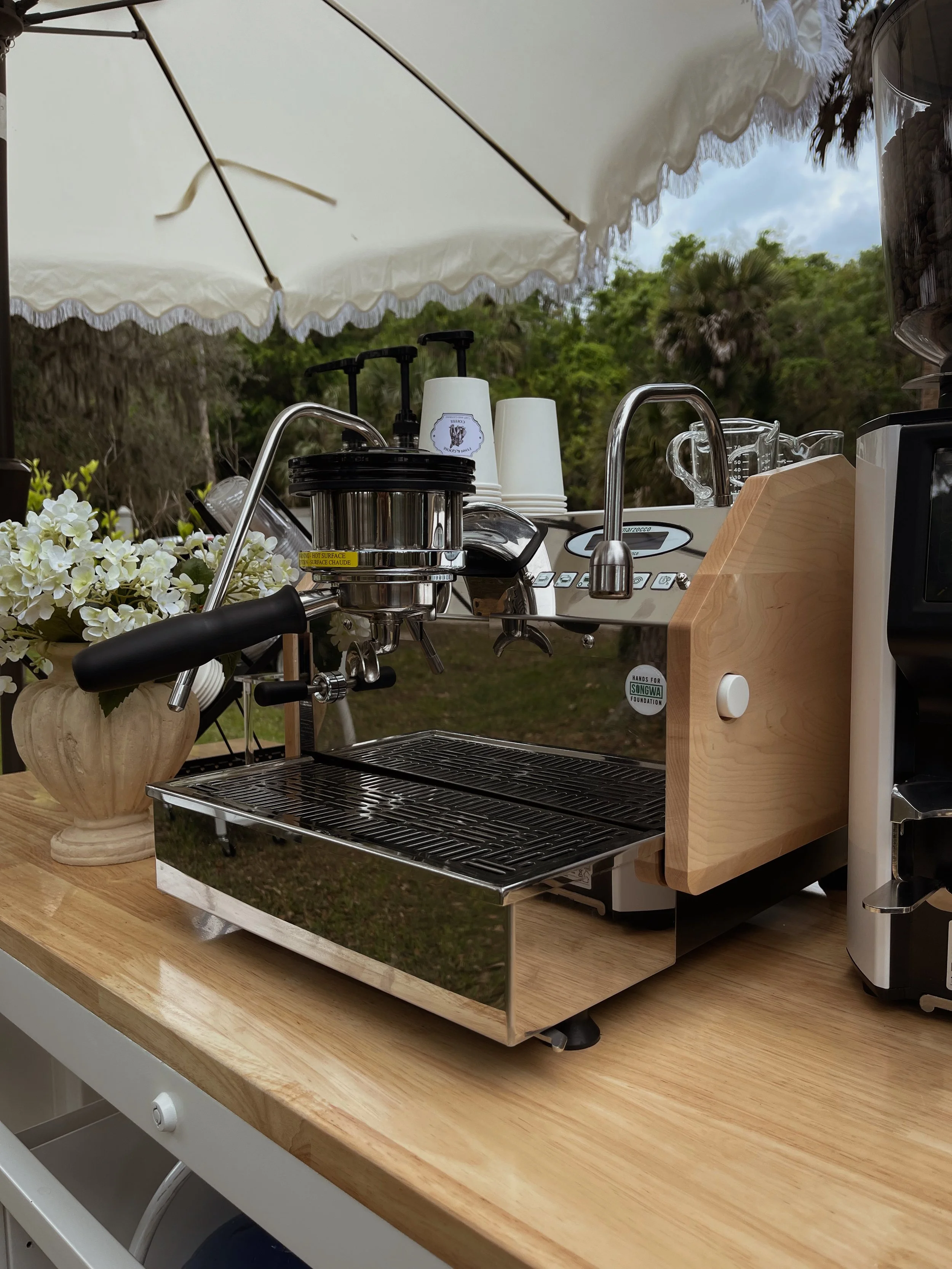 Coffee machine on a wooden countertop with cups, flowers, and an outdoor umbrella.