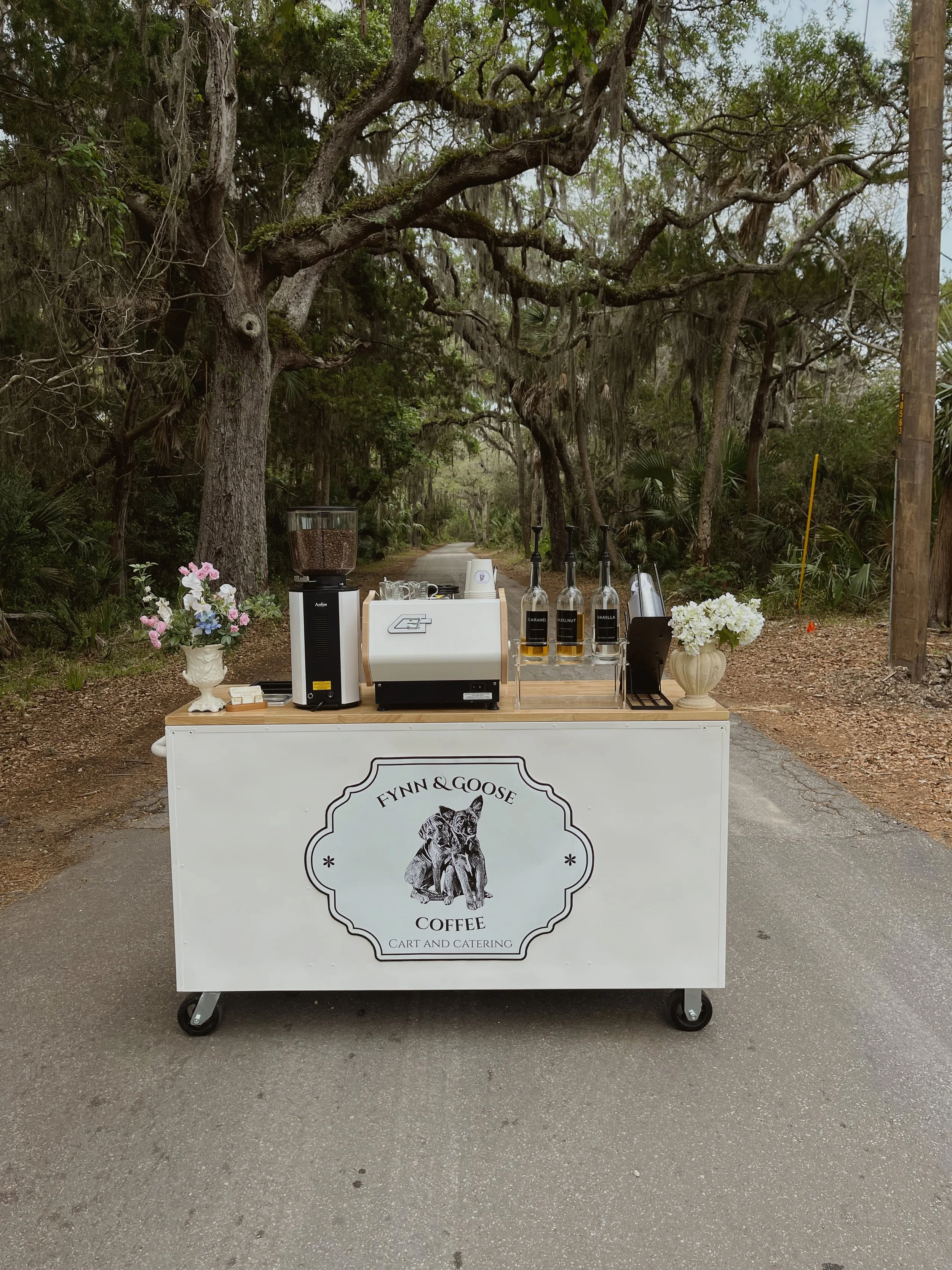 A white coffee cart with a logo featuring a wolf, offering coffee and cart catering, set up outdoors on a road with trees in the background.