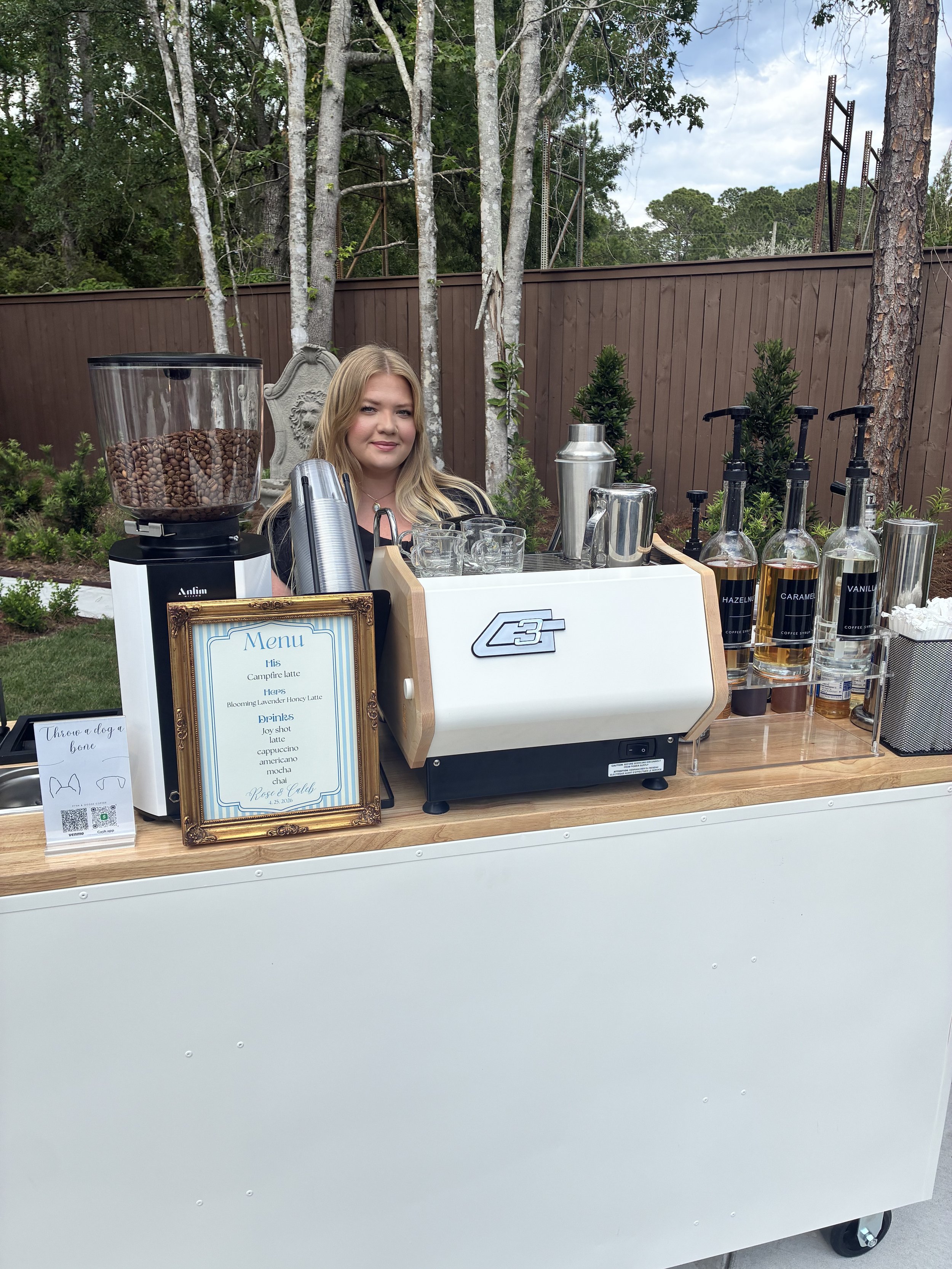 A young woman standing behind a coffee bar outdoors, with various coffee-making supplies and bottles of flavored syrup on the counter, trees and a brown wooden fence in the background.