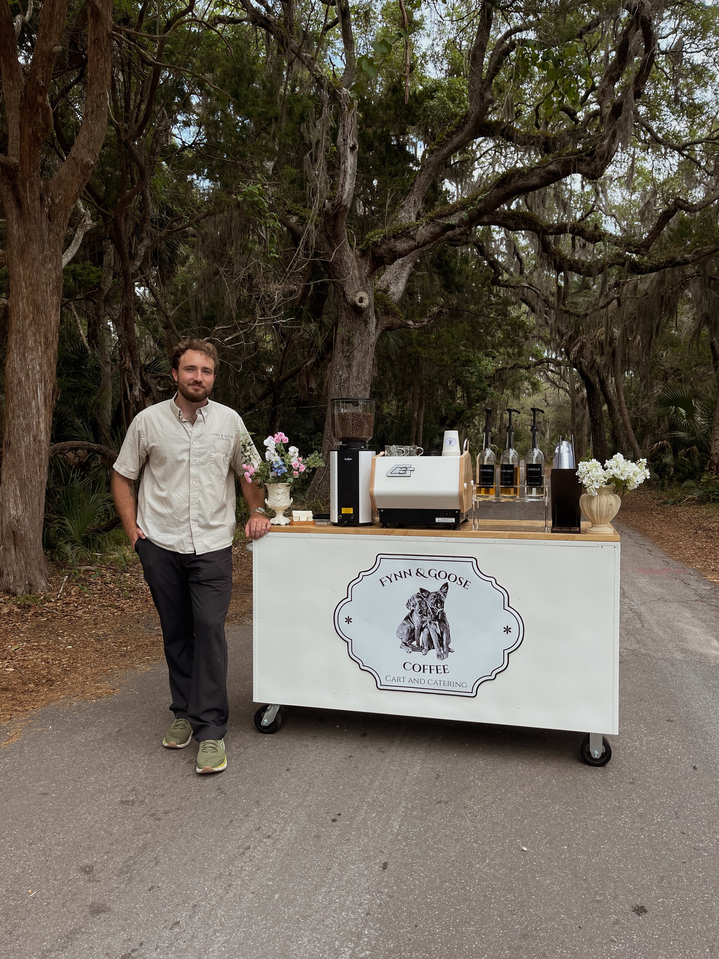 A man standing next to a coffee cart labeled "Fynn & Goose Coffee Cart and Catering," set outdoors with trees in the background. The cart has coffee brewing equipment, cups, and decorative flower arrangements.