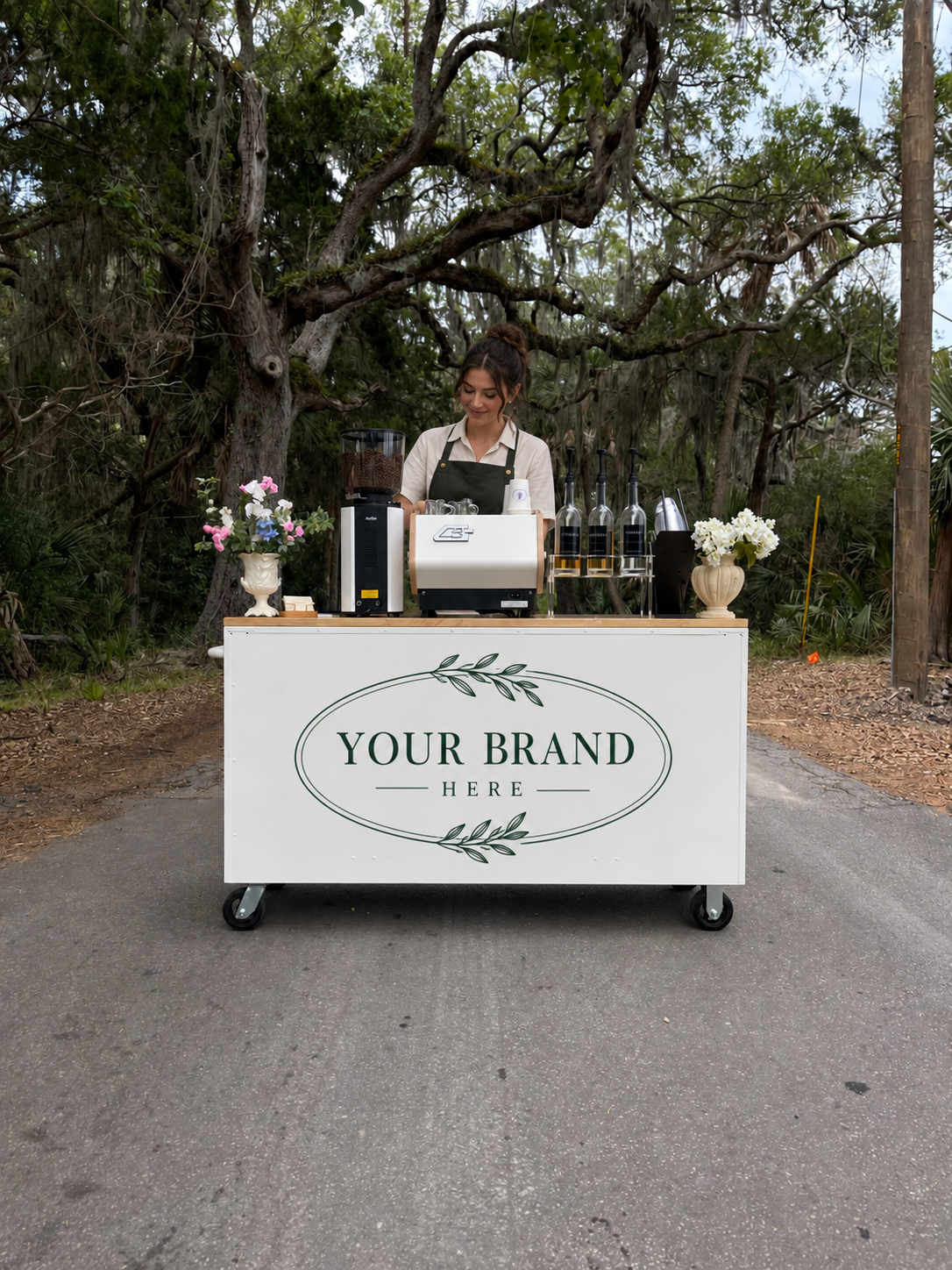 Outdoor mobile coffee bar with a barista, espresso machine, coffee grinder, floral arrangements, and customizable “Your Brand Here” signage in a lush garden setting
