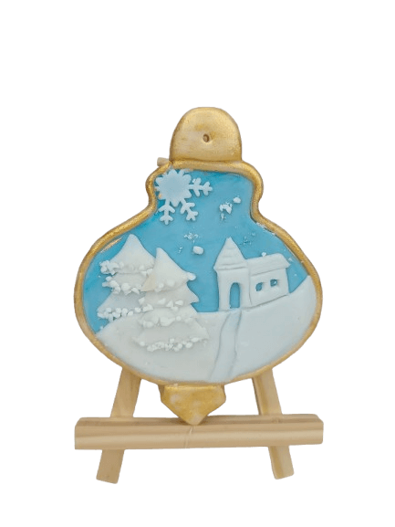 Decorated gingerbread cookie shaped like a snow globe with a winter scene, including snow-covered trees, a small house, and snowflakes, on a wooden stand.
