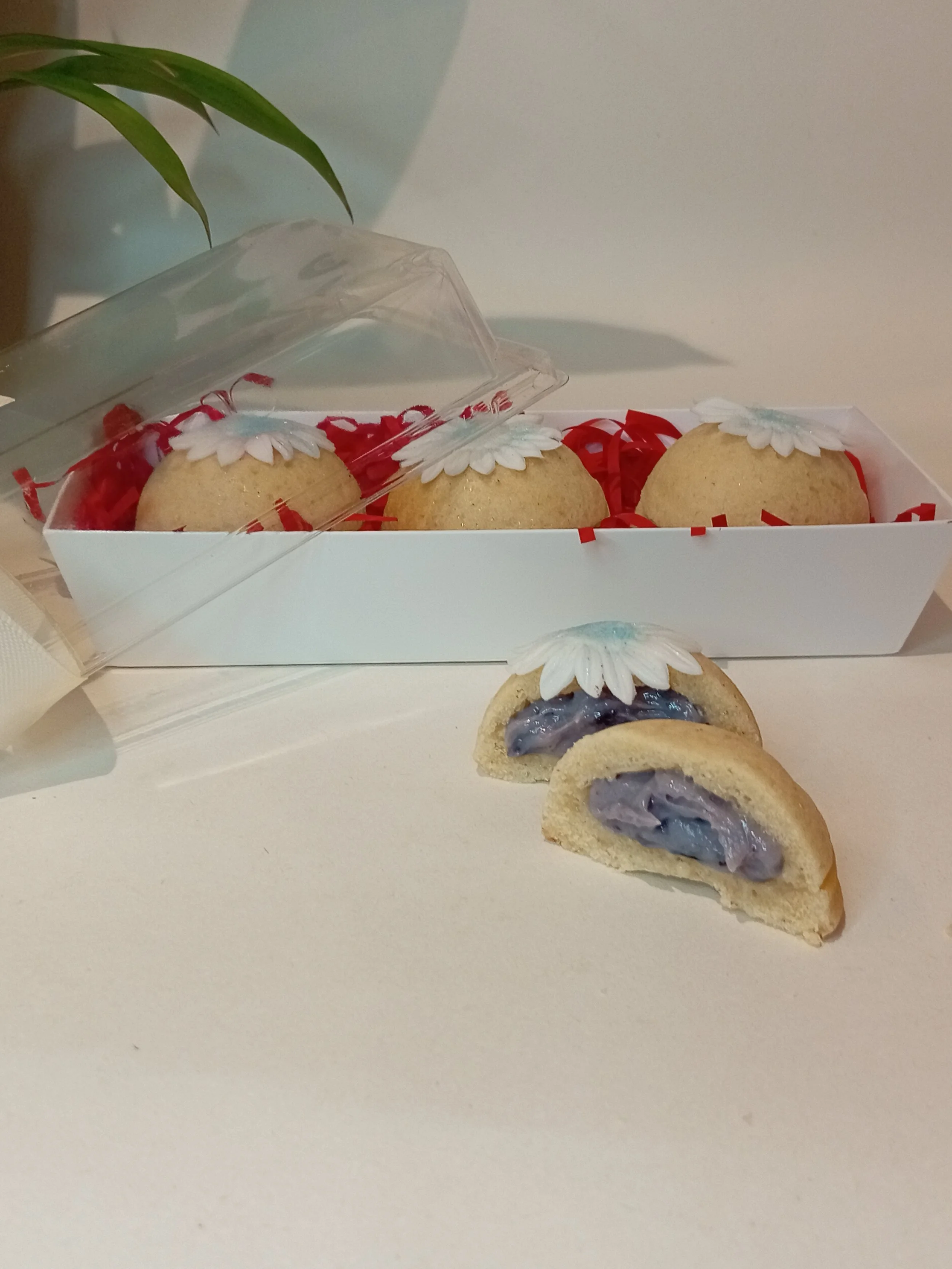 A box of three decorated cookies with white flower designs on top, partially open, with two of the cookies cut in half revealing a purple filling inside.
