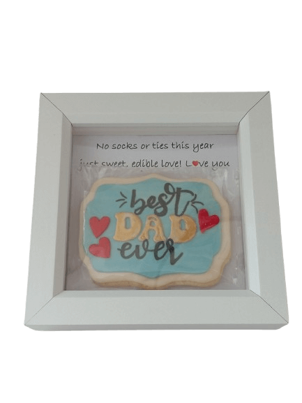 A decorated cookie in a white frame with a handwritten note behind it. The cookie says 'best DAD ever' with hearts on a blue background.