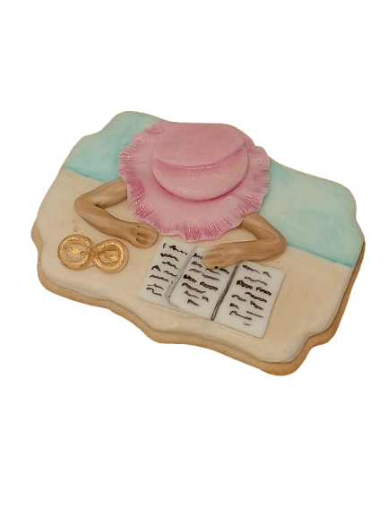 Decorative cookie in shape of a person reading a book, wearing a pink hat with a ruffled edge, lying on a blue and white surface.