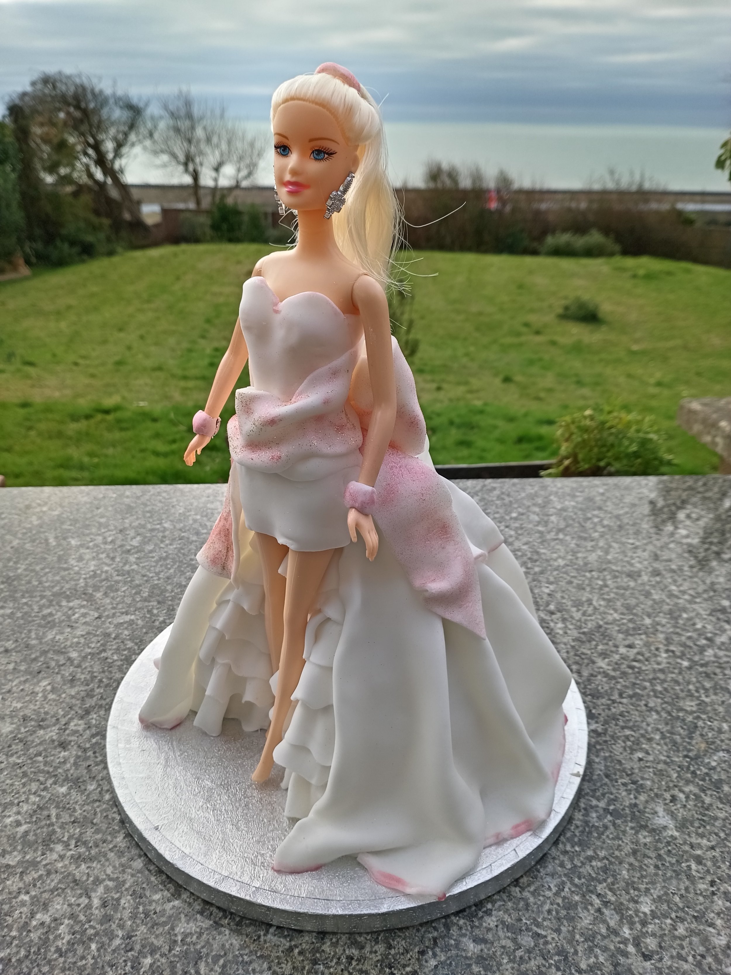 A Barbie doll dressed in a pink and white gown resembling a wedding dress, standing outdoors on a table with a grassy field and cloudy sky in the background.