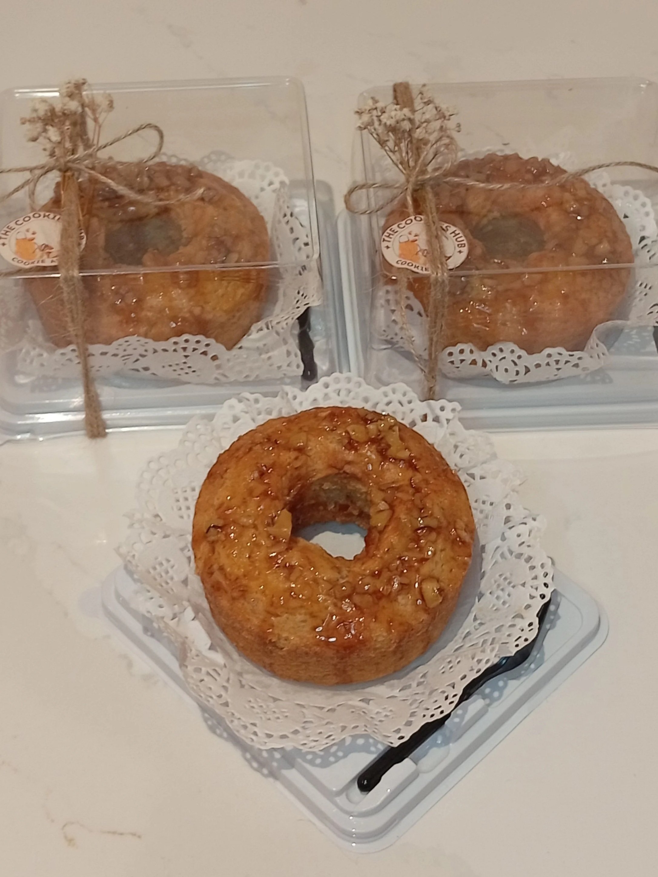 Three glazed donuts with chopped nuts, one in front and two in boxes behind, on a white surface.