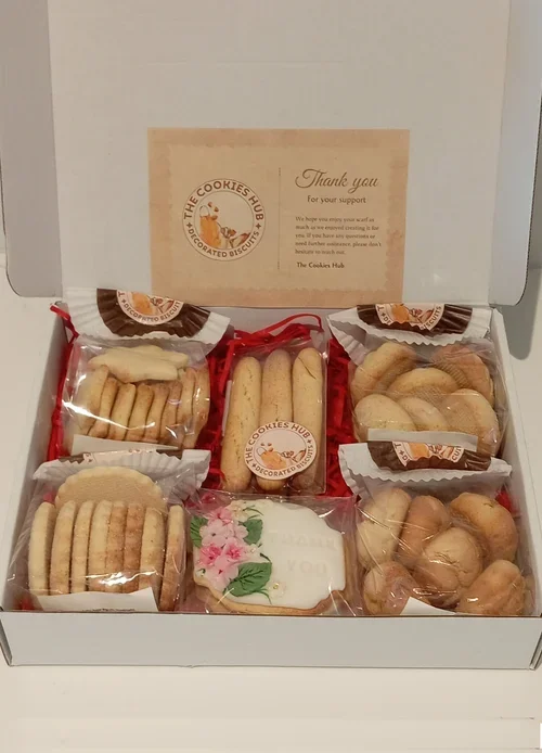 An open white cookie box containing assorted cookies, some in plastic wrapping, with a thank you note inside the lid.