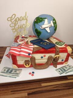 A cake designed like a suitcase with a world map, a small globe with an airplane, and travel-themed decorations including a 'Good Luck' topper and dollar bills.