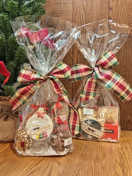 Two clear plastic gift bags tied with plaid ribbons, containing holiday cookies and baked goods, placed on a wooden surface beside a decorated Christmas tree.
