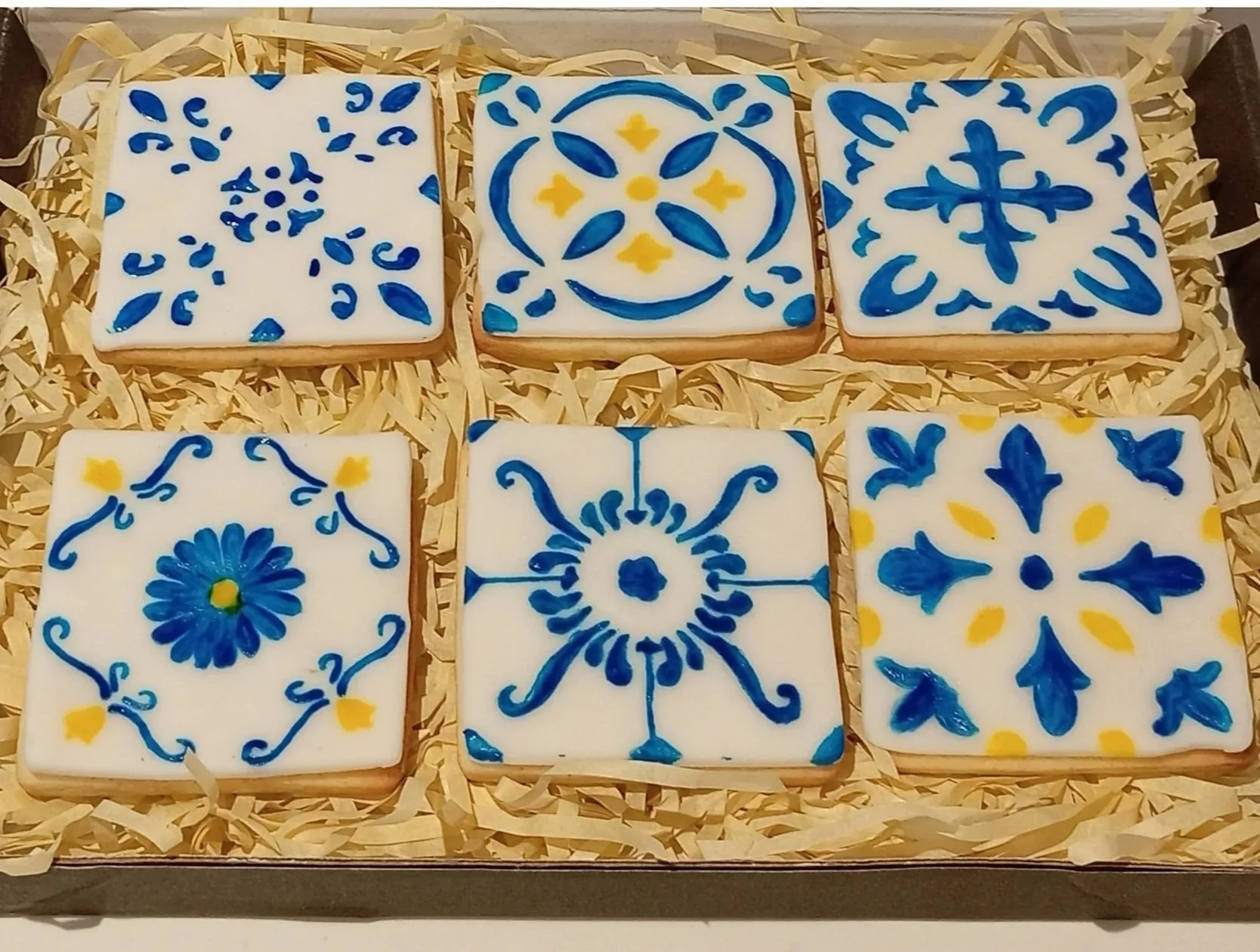 Six tiles with blue and yellow decorative patterns on a white background arranged in two rows of three on shredded paper.