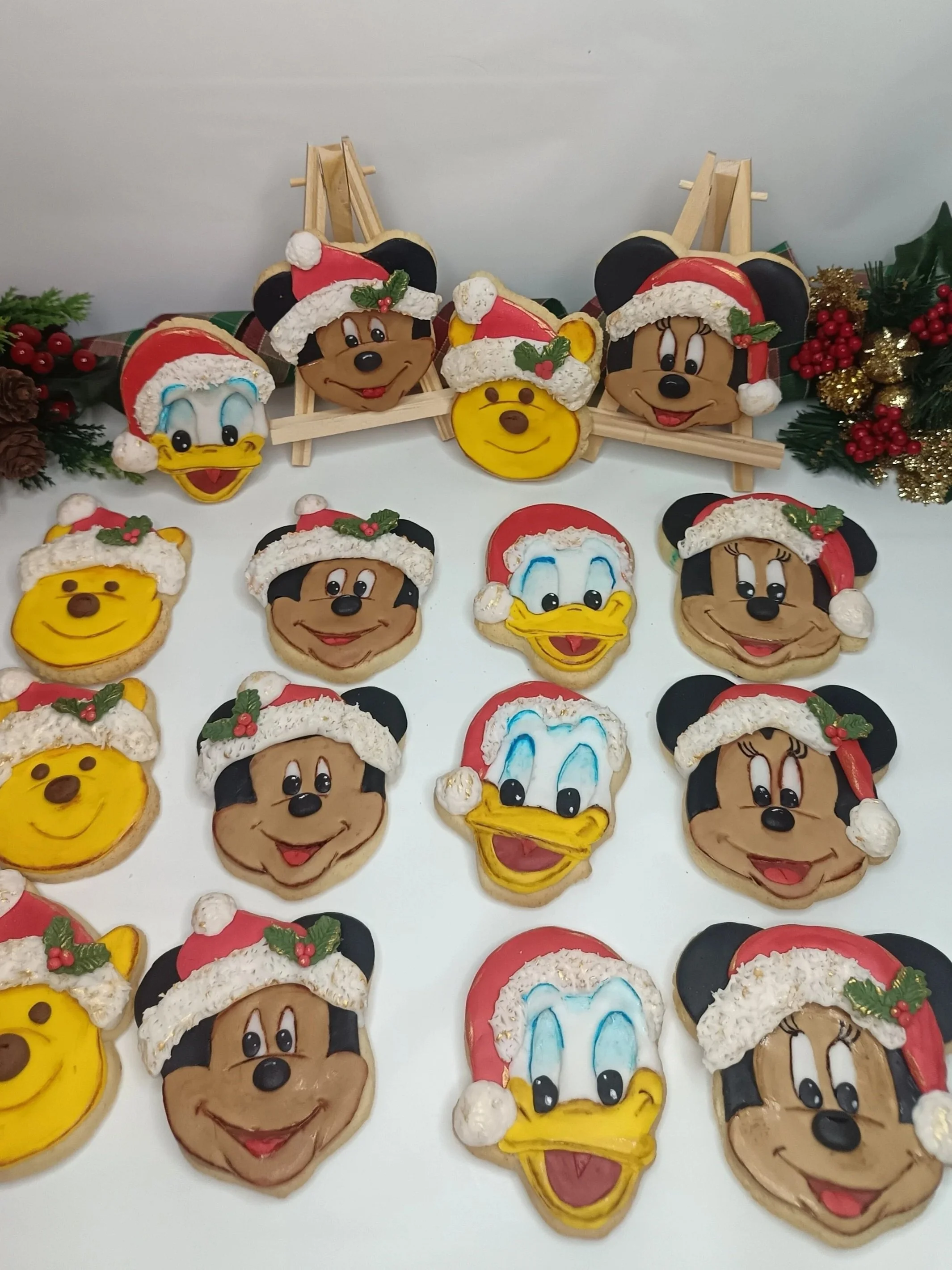 Christmas decorated cookies shaped like Disney characters Mickey Mouse, Minnie Mouse, Donald Duck, and Pluto, all wearing Santa hats, arranged on a table with holiday decorations.