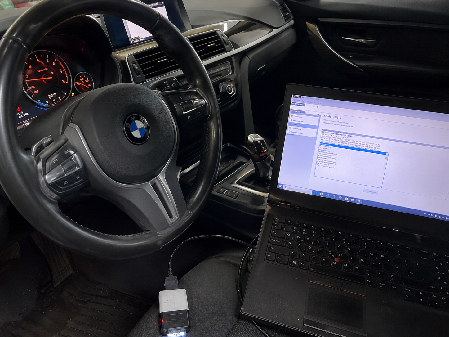 The interior of a BMW vehicle with a steering wheel, dashboard, gear shift, and a laptop on the passenger seat showing a software interface.