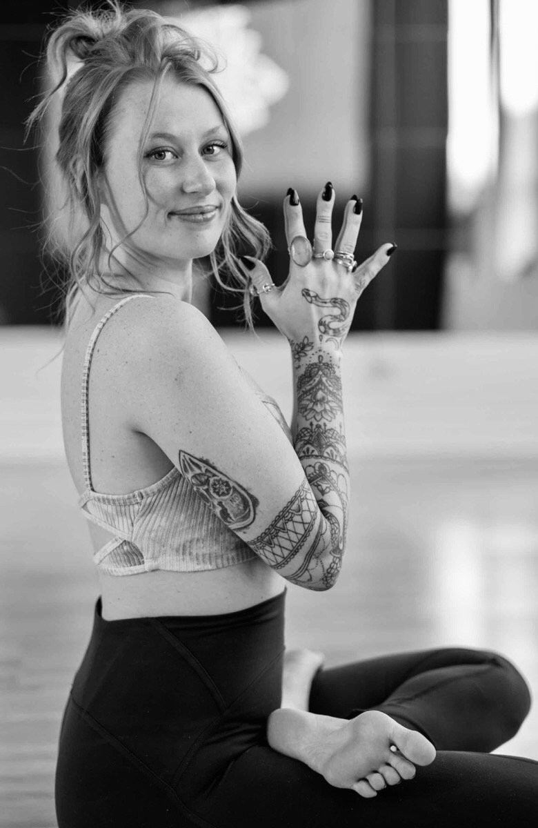 Woman with tattoos on her arms and hand, sitting cross-legged on the floor, smiling and looking at the camera, in an indoor setting.