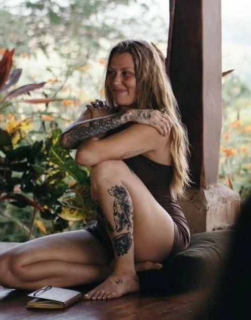 A woman sitting on a wooden deck, hugging her knees, with tattoos on her arm and leg, smiling and looking to the side. She is in a natural setting with greenery in the background.