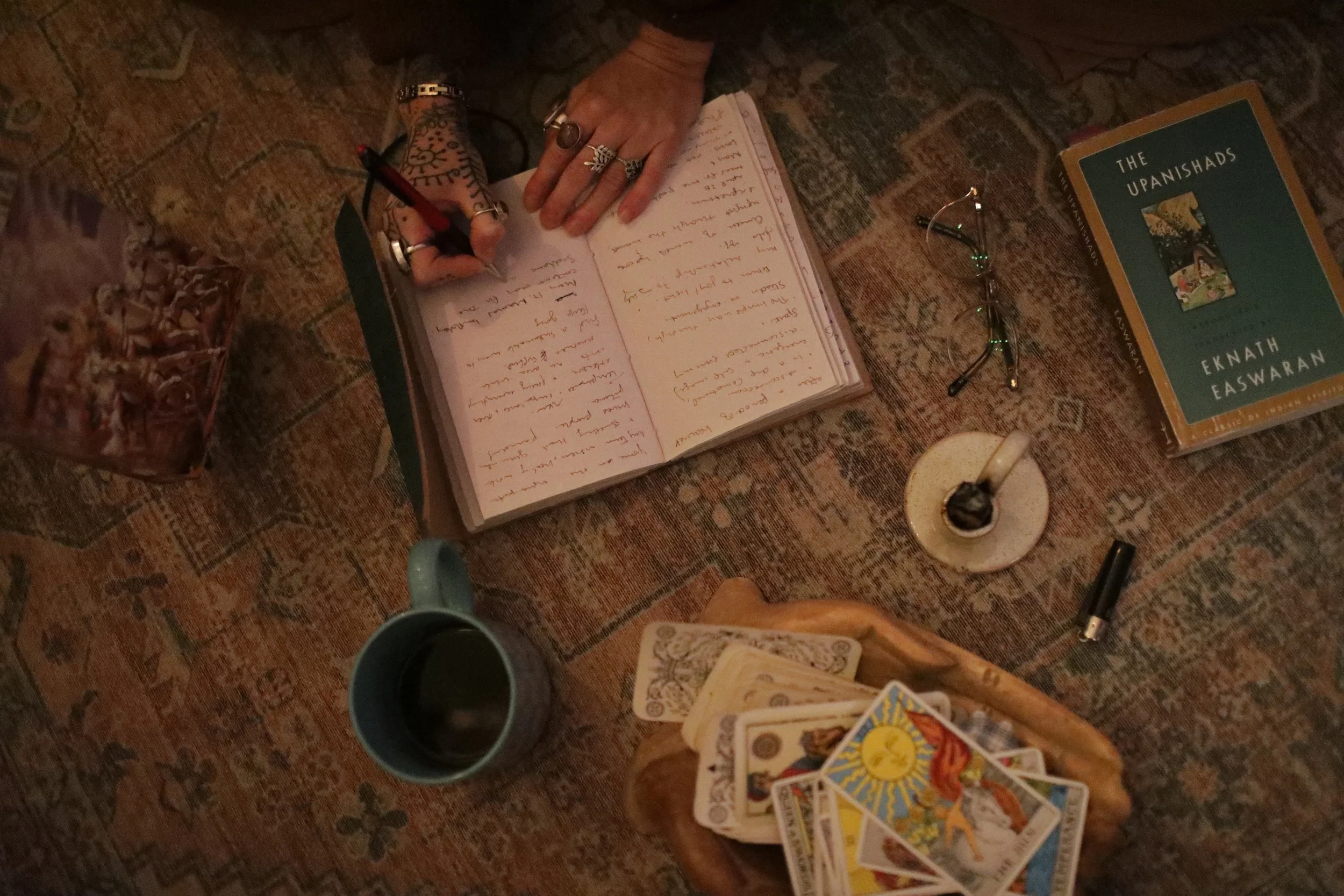 A person with henna tattoos on their hand writes in a notebook on a patterned rug. Nearby are reading glasses, a book titled 'The Upanishads,' a cup of coffee, tarot cards, and a deck of cards.