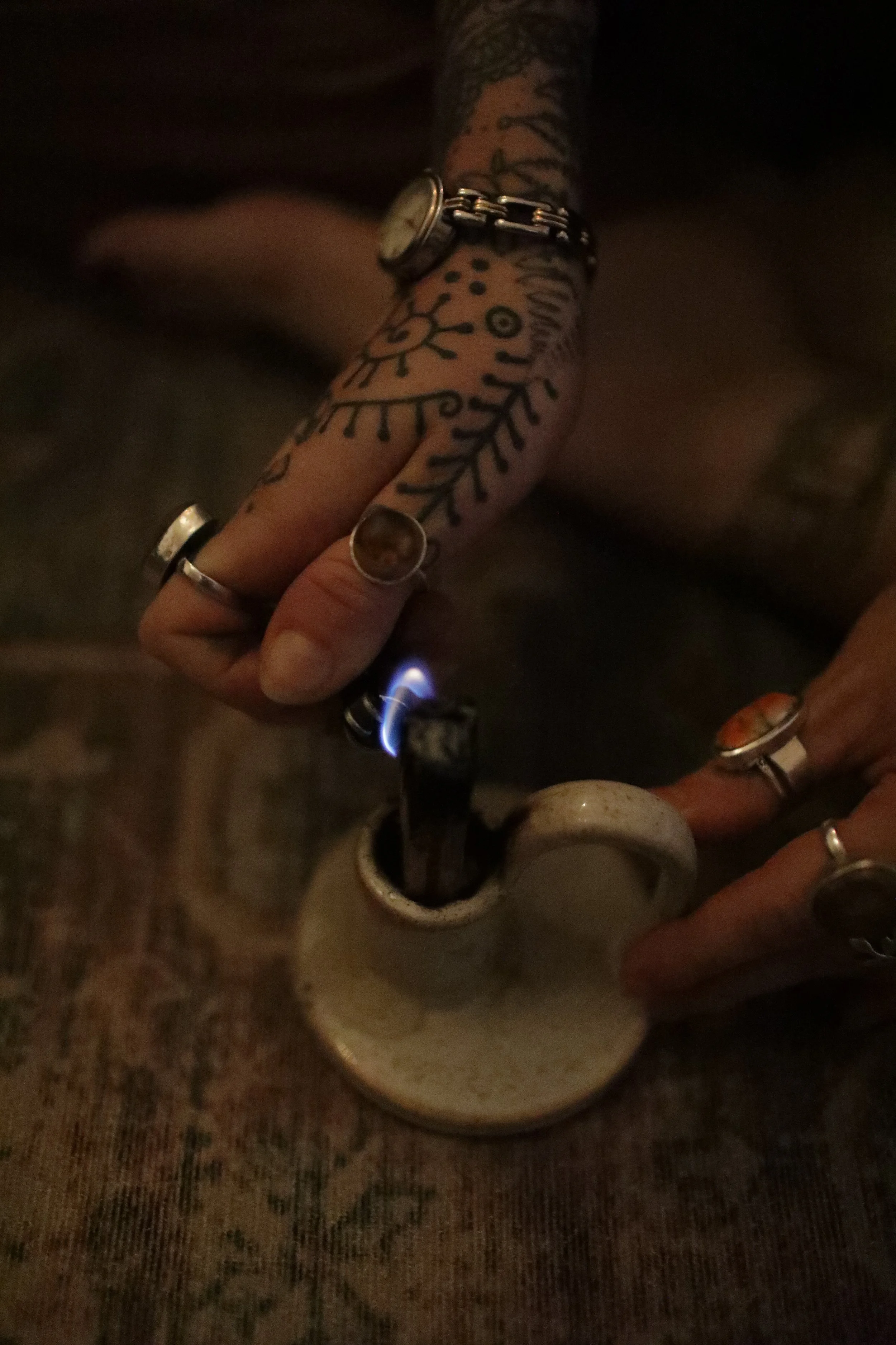 A person's tattooed hand with rings is holding a lighter over a mug of coffee, igniting a flame, in a dimly lit setting.