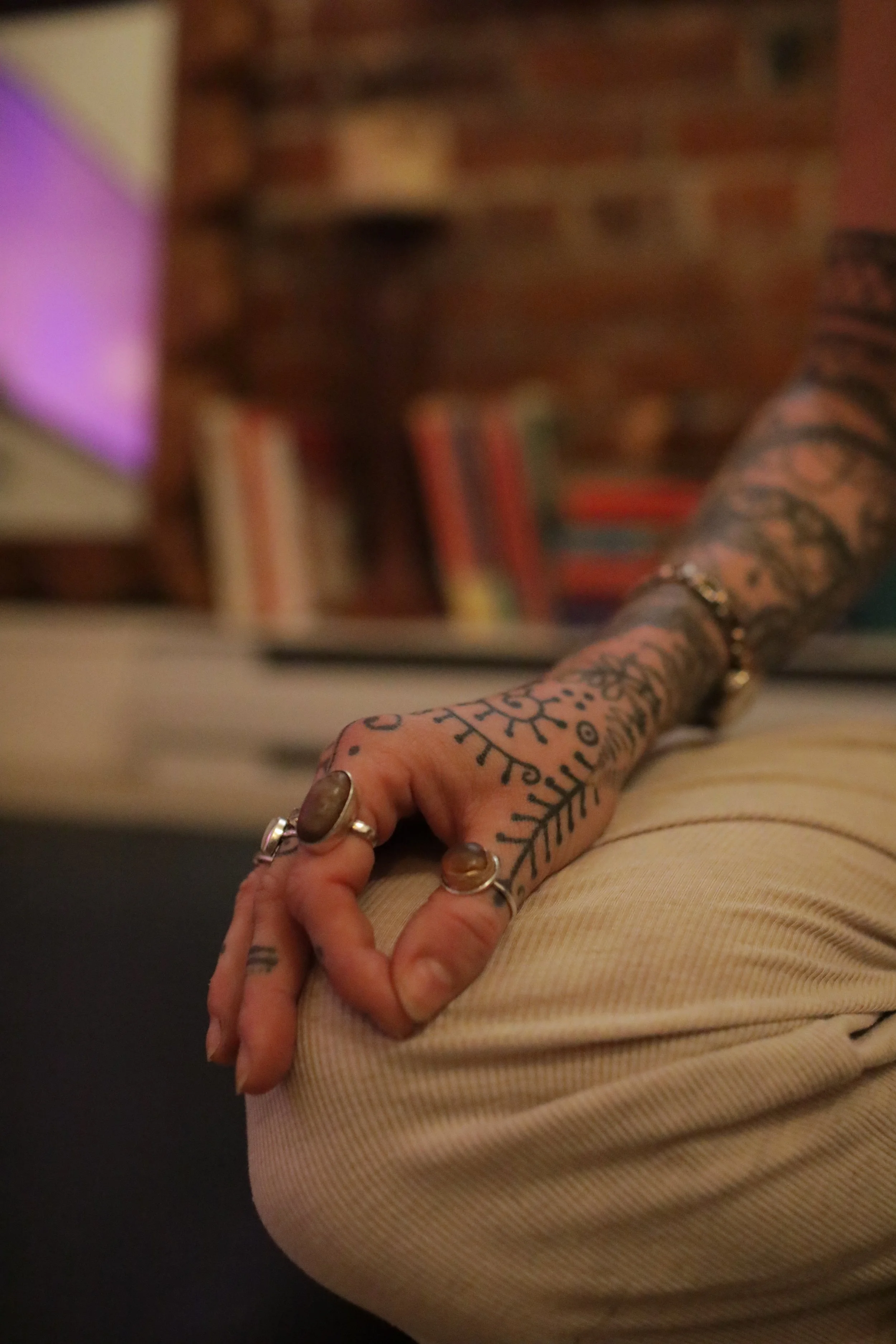 Close-up of a person's arm with tattoos, wearing rings, resting on their knee, with a bookshelf in the background.