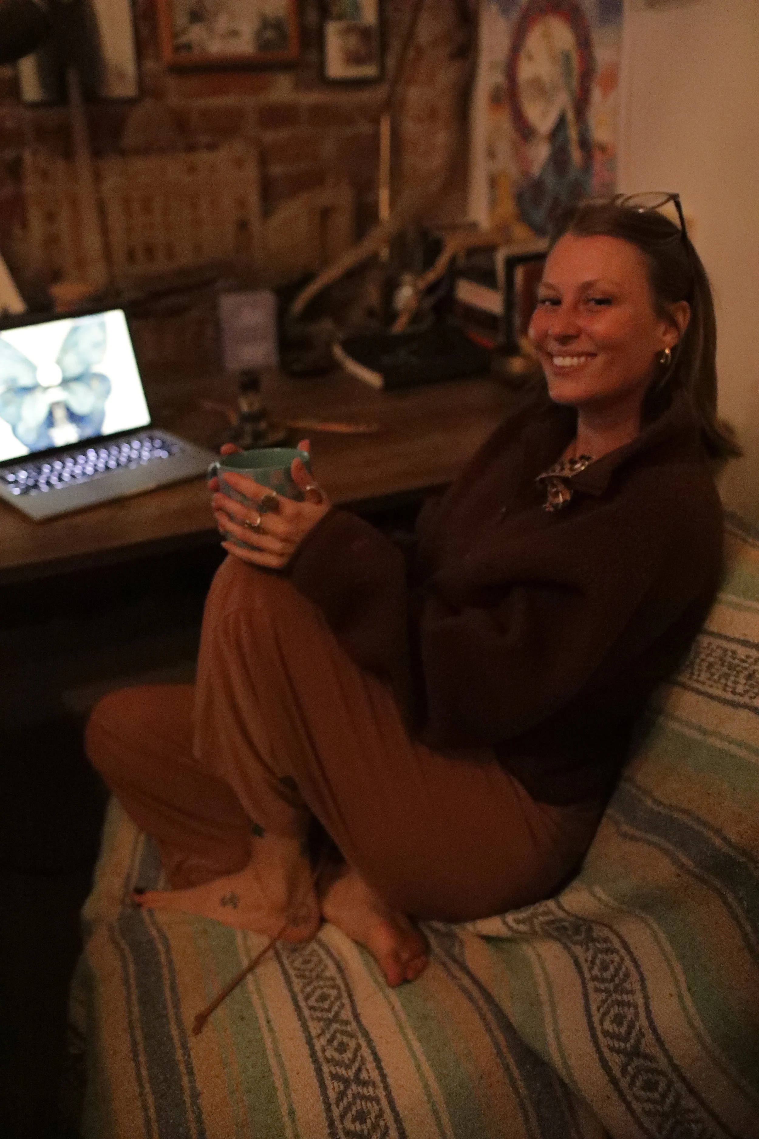 A woman with glasses on her head, smiling, sitting cross-legged on a patterned rug in a room with brick walls, holding a mug, with various pictures and artwork on the wall behind her.