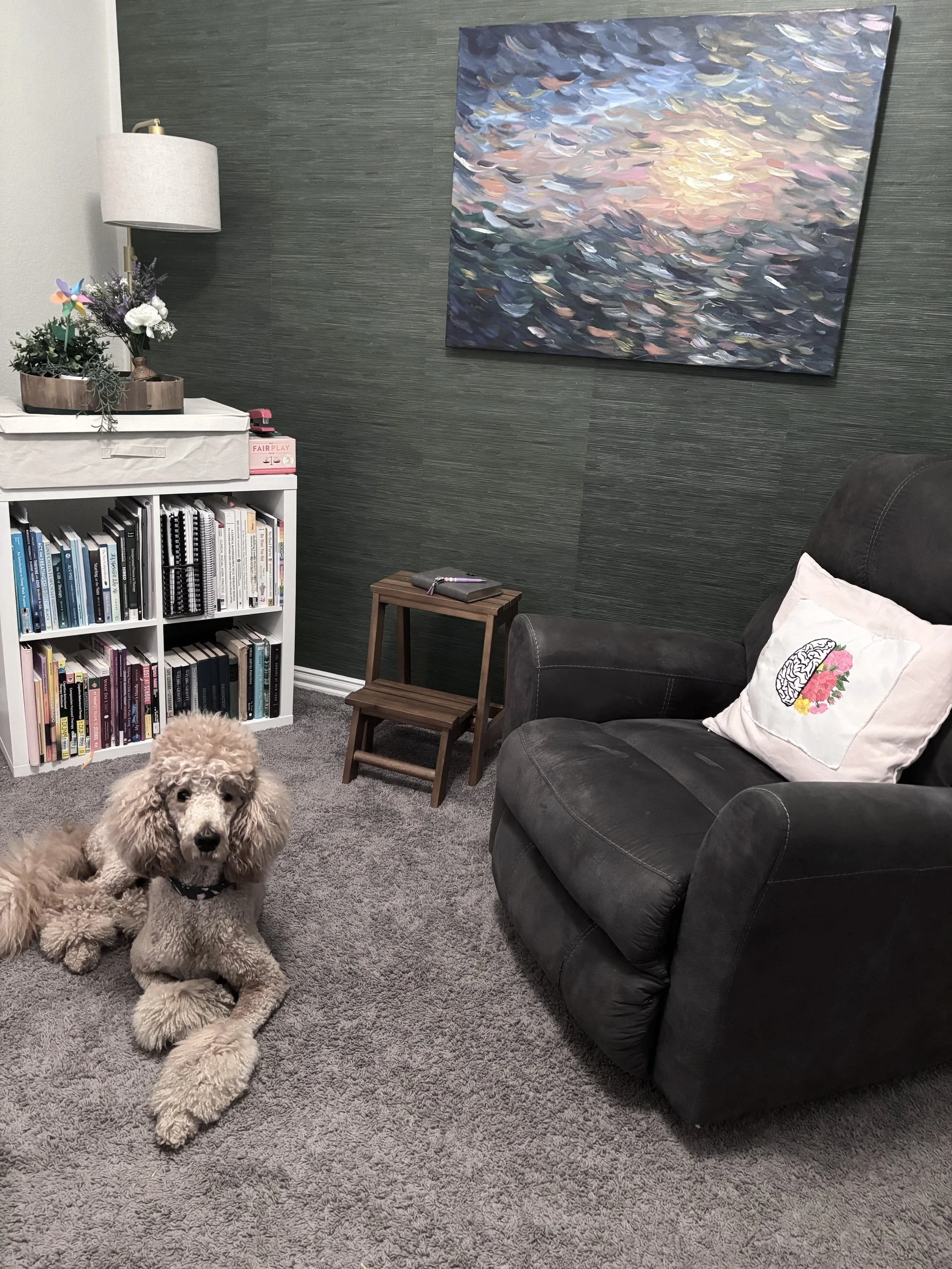 A cozy living room corner featuring a black armchair with a decorative pillow, a small wooden side table, a lamp, a bookshelf filled with books, a large abstract ocean painting on the wall, and two poodles sitting on a grey carpet.