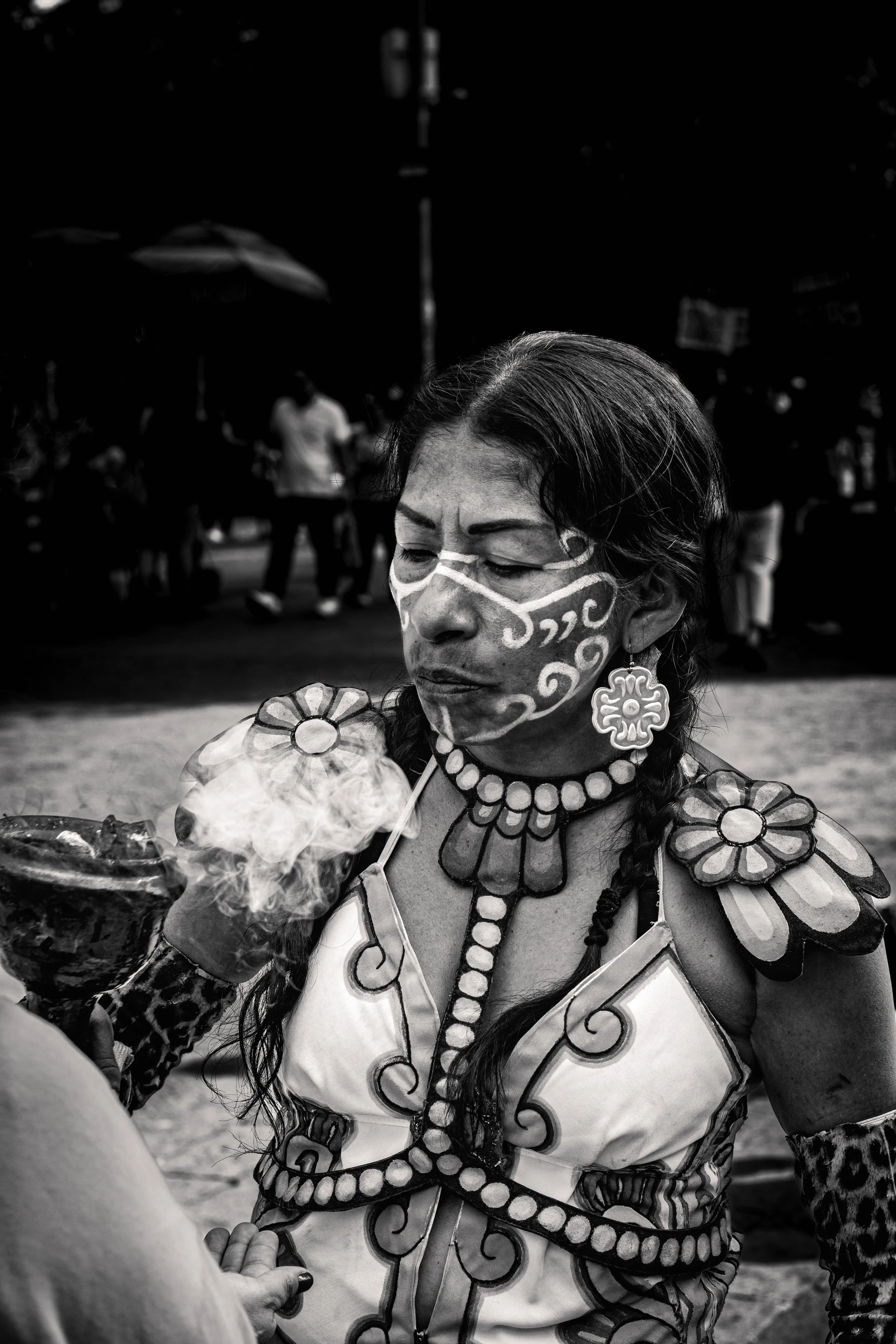 A woman dressed in traditional indigenous attire with face paint, wearing floral earrings and necklaces, holds a bowl with smoke rising from it during a cultural festival or ceremony.