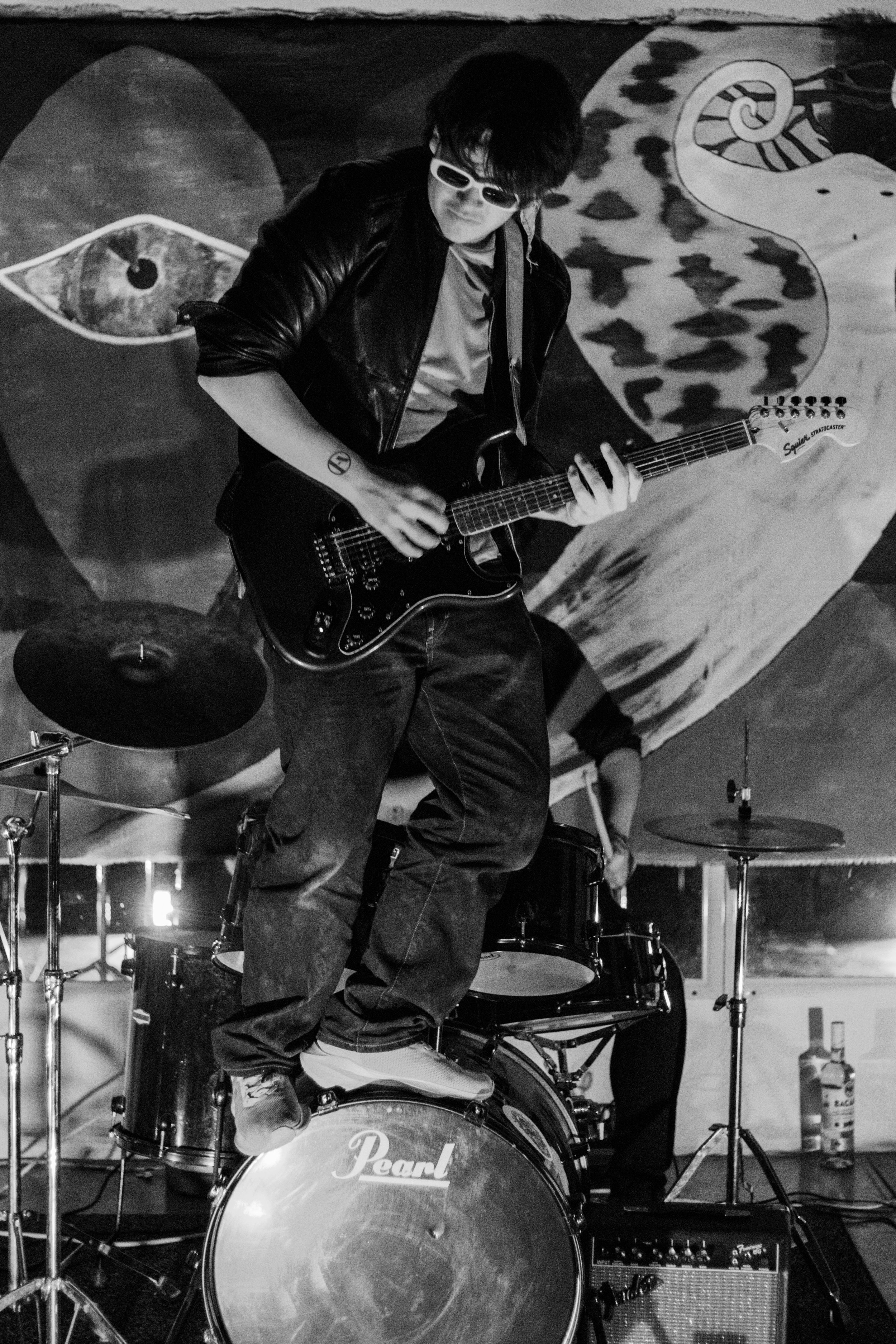 A person playing electric guitar standing on a drum set in a music venue with a large eye and animal artwork mural in the background, in black and white.