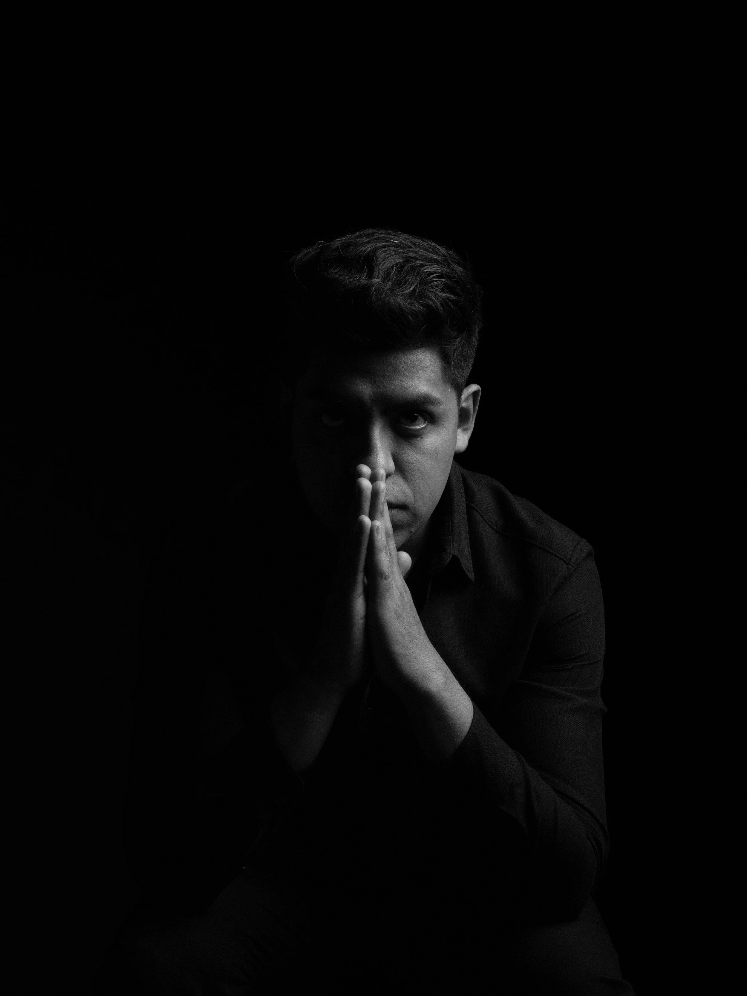 A young man with dark hair, dressed in a black shirt, sits with his hands pressed together near his face, looking intently at the camera in a dark setting with low-key lighting.