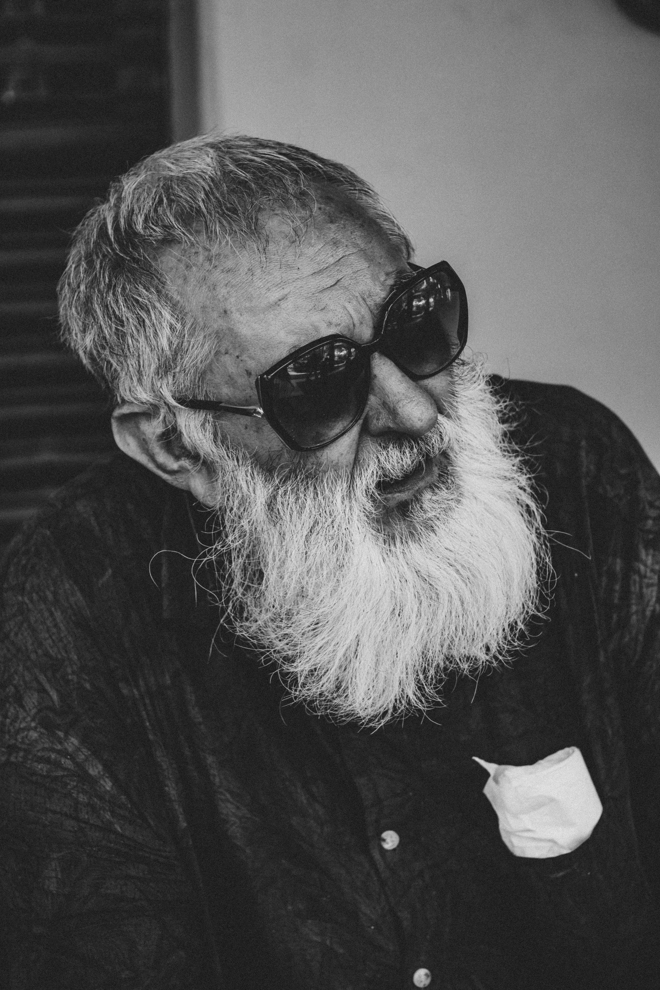 Black and white photo of an elderly man with a long white beard wearing large dark sunglasses and a dark shirt. A tissue is tucked into his shirt pocket.