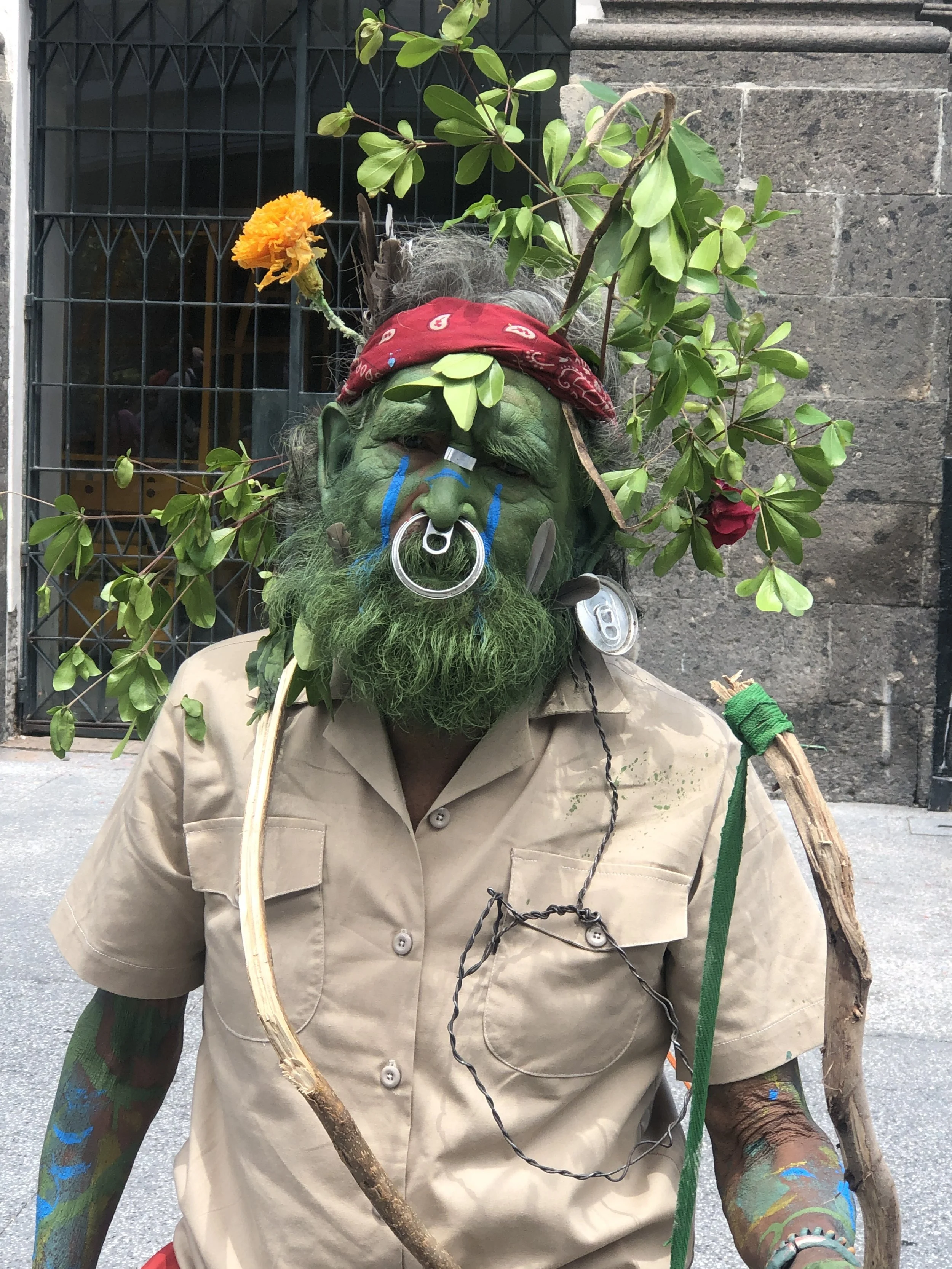 Person dressed as a green-skinned forest or nature character with leaves and flowers, wearing a tan uniform shirt and a red bandana, with face paint, a nose ring, and painted arms.