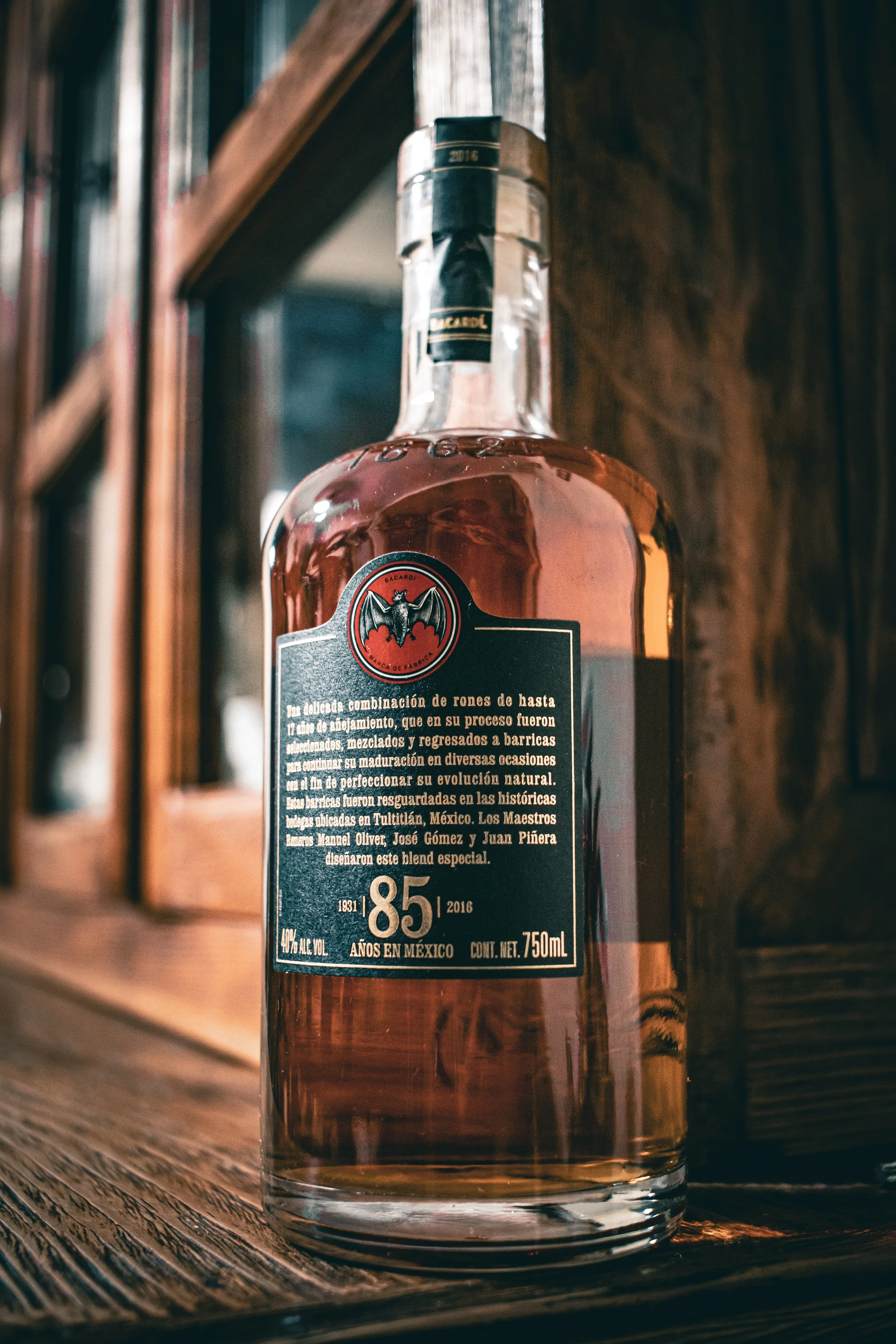 A bottle of Rum Bacardi 85 years in Mexico, with a black label featuring a red and black bat logo, placed on a wooden surface against a wooden background.