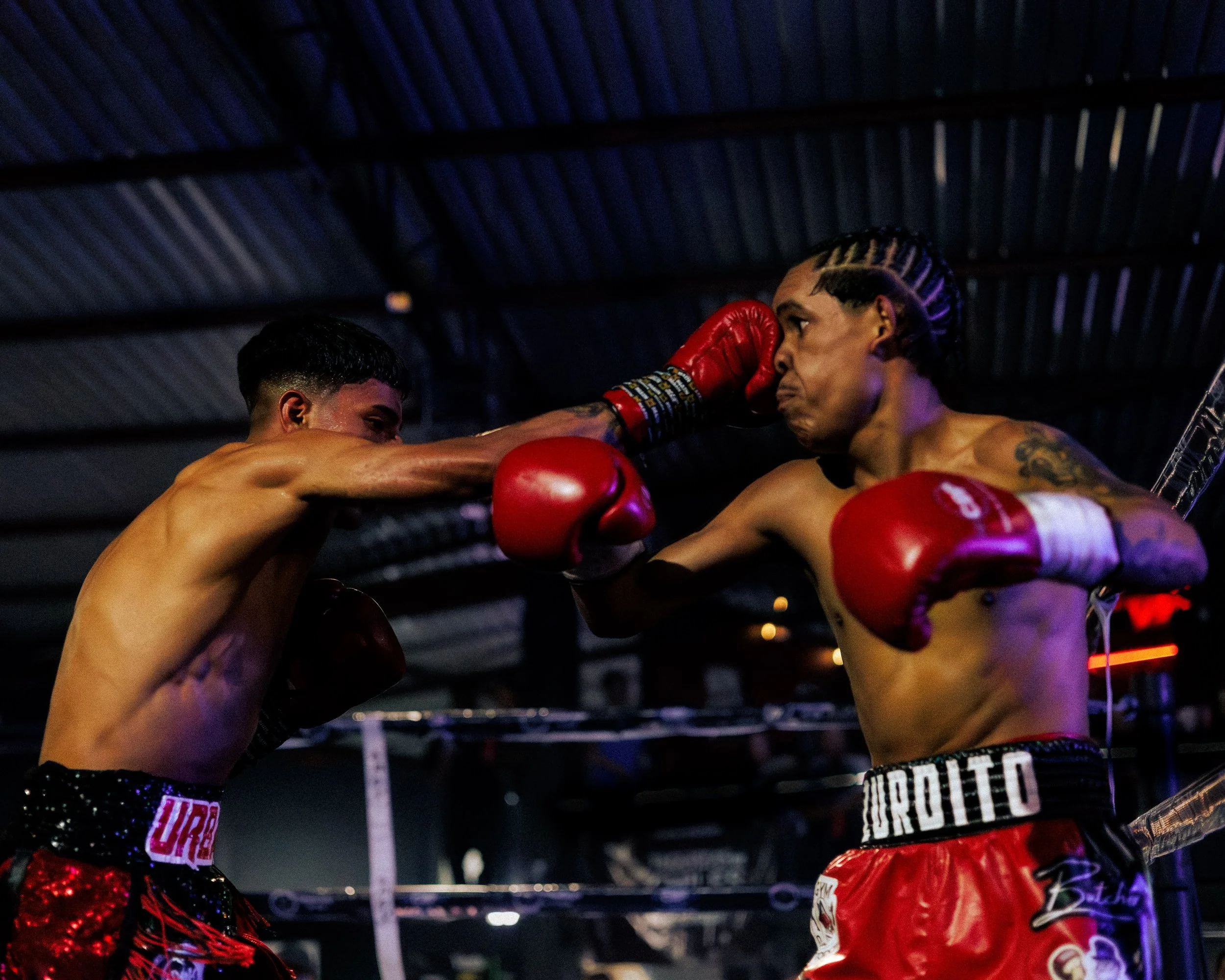 Two male boxers in a ring, one delivering a punch to the other's face under low lighting.