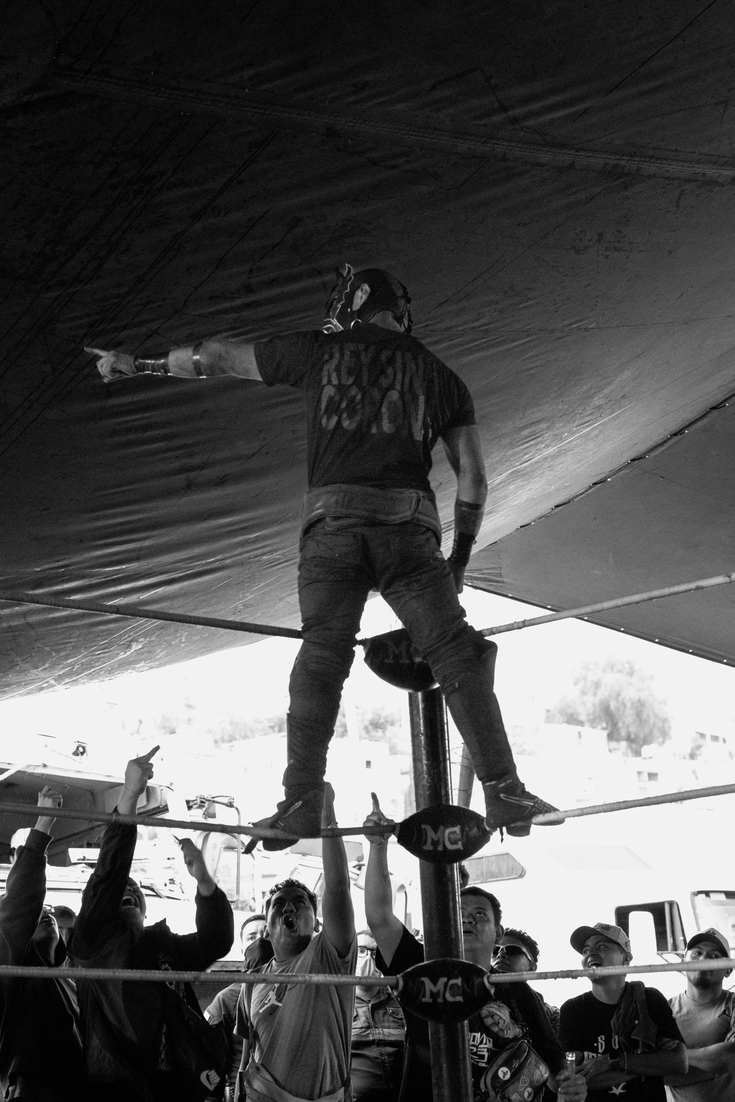 A performer stands on a tightrope, pointing with one arm while wearing a bandana and a T-shirt with the word 'Rey' visible. Several people below watch and cheer during an outdoor event.