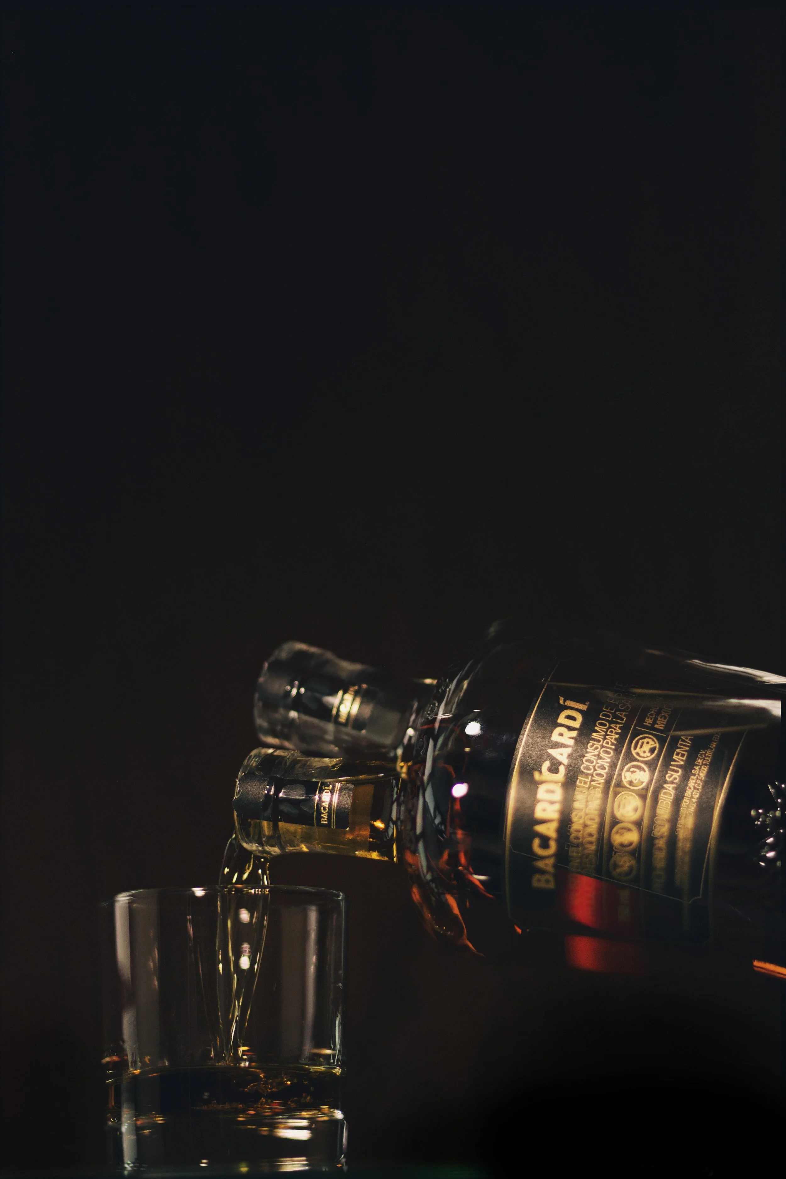 A bottle of Bacardi rum pouring into a glass against a dark background.