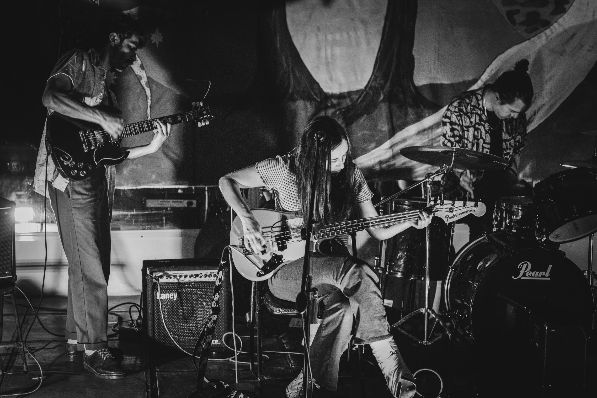 Three musicians performing on stage: a male guitarist on the left, a female bassist in the center, and a male drummer on the right, with a painted backdrop.