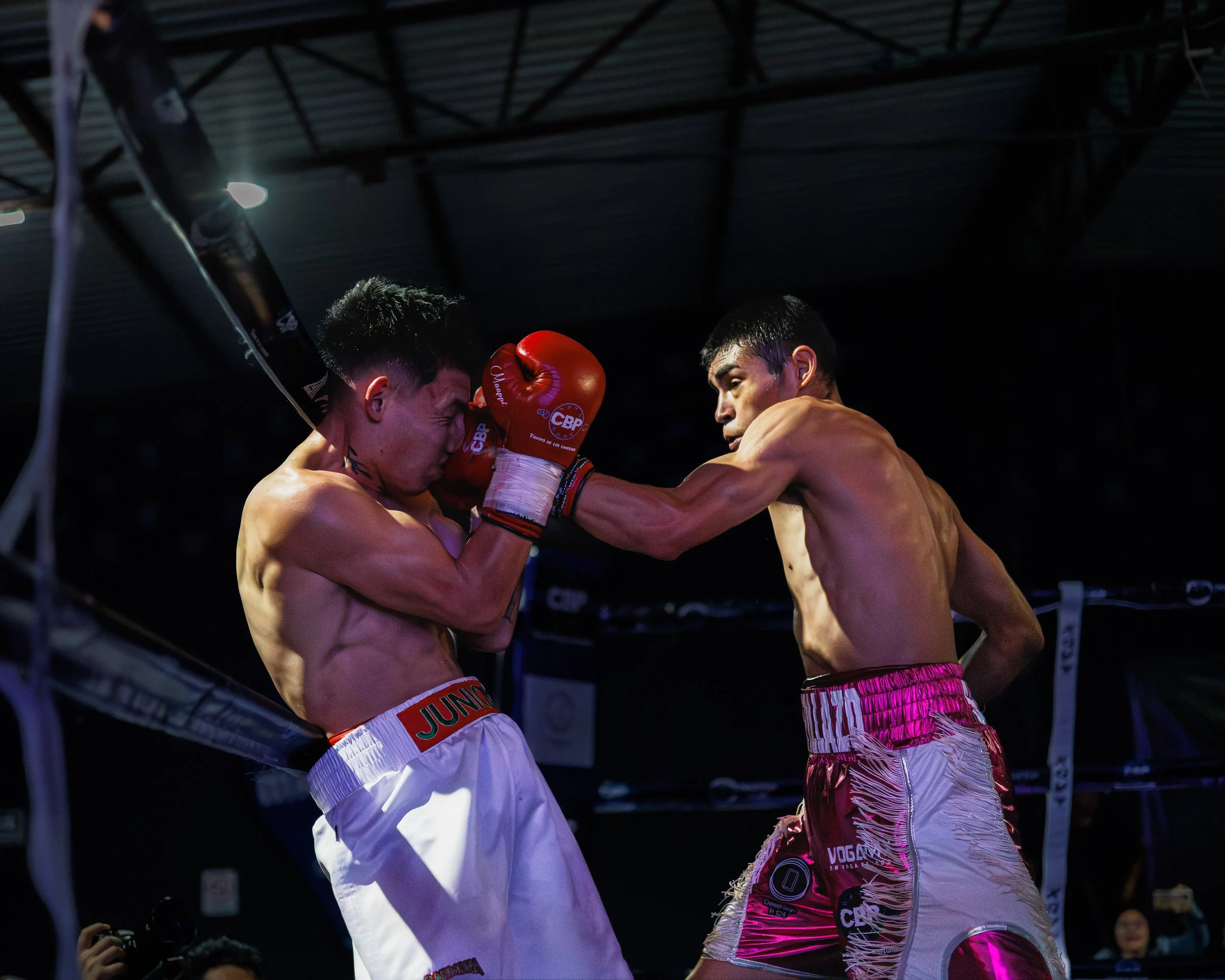 Two shirtless male fighters in a boxing ring, one delivering a punch to the other's face, wearing red boxing gloves and shorts with branding, under overhead lighting