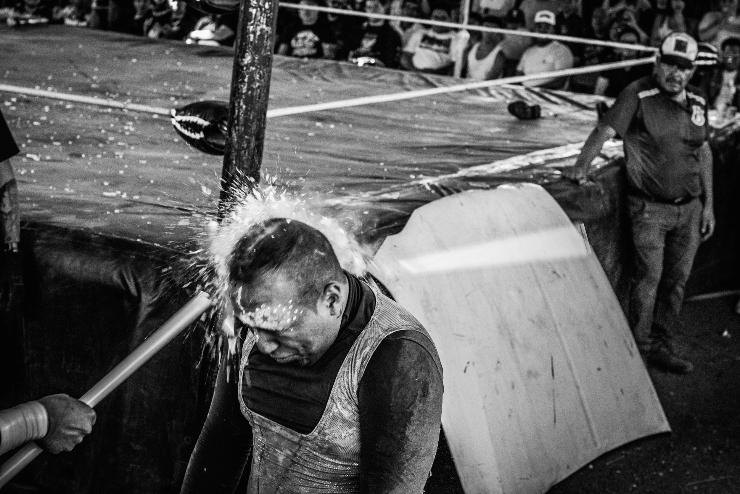 A man getting hit on the head with a pole, water splashing over him, as people stand and watch in the background.