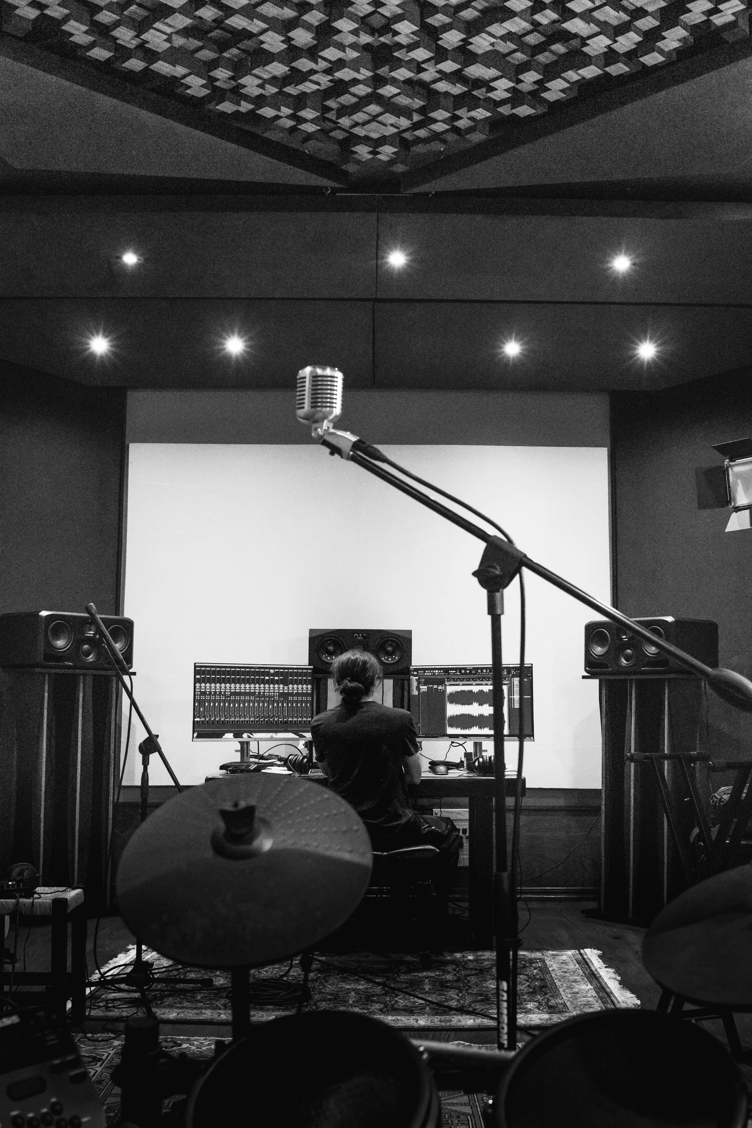 A black and white photo of a music studio with a person working on a computer with studio speakers, microphones, and a drum set in the foreground.