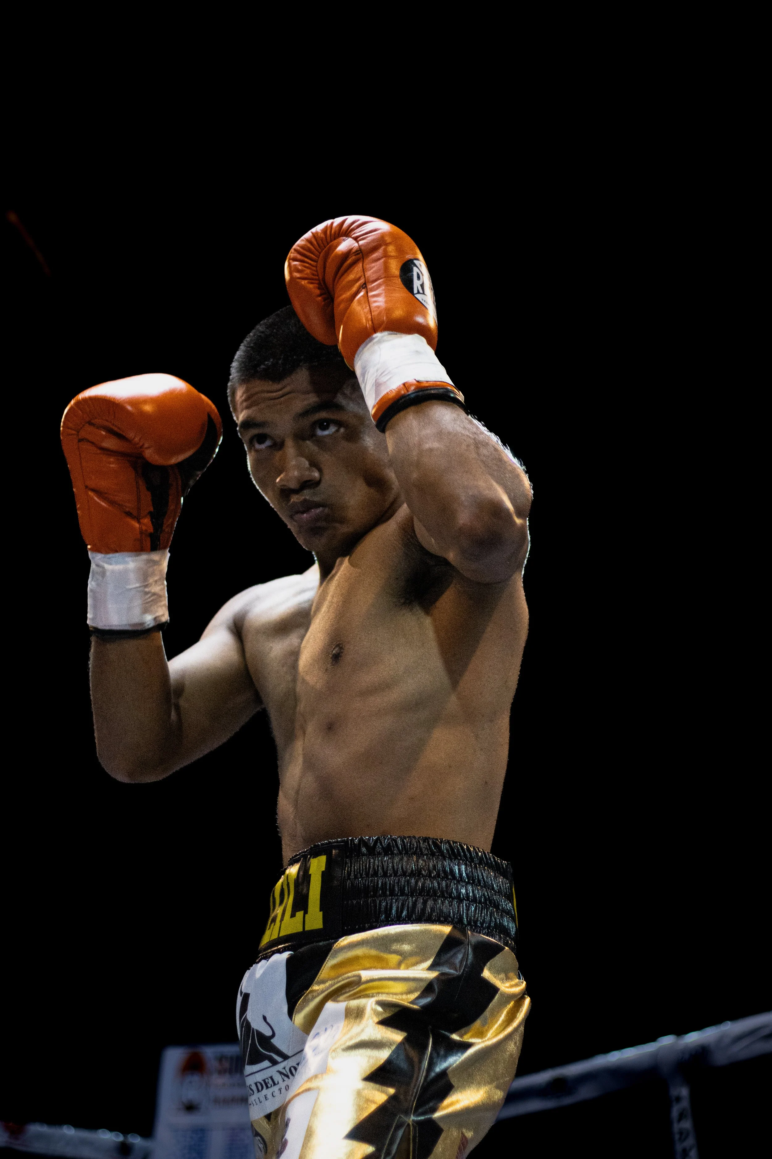 A shirtless male boxer wearing orange gloves, black and gold boxing shorts, preparing for a fight, standing in a boxing ring with a dark background.
