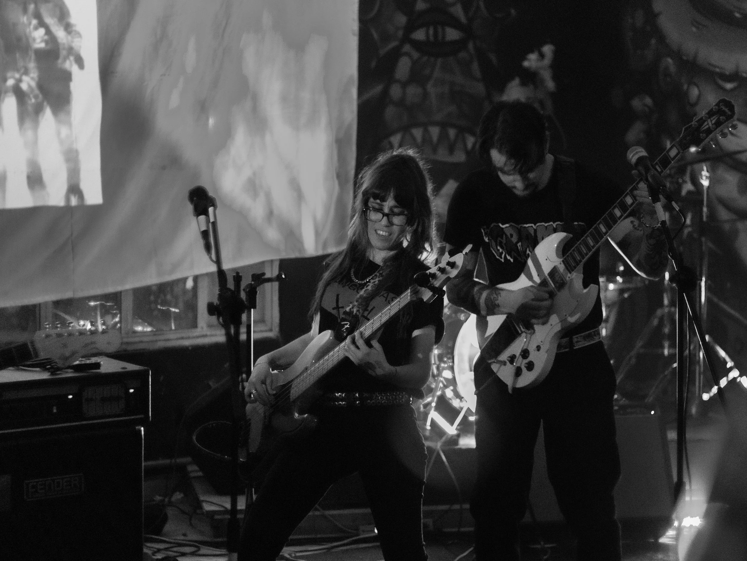 Two musicians, a woman and a man, playing guitars on stage during a live performance, with large artwork or projection in the background.
