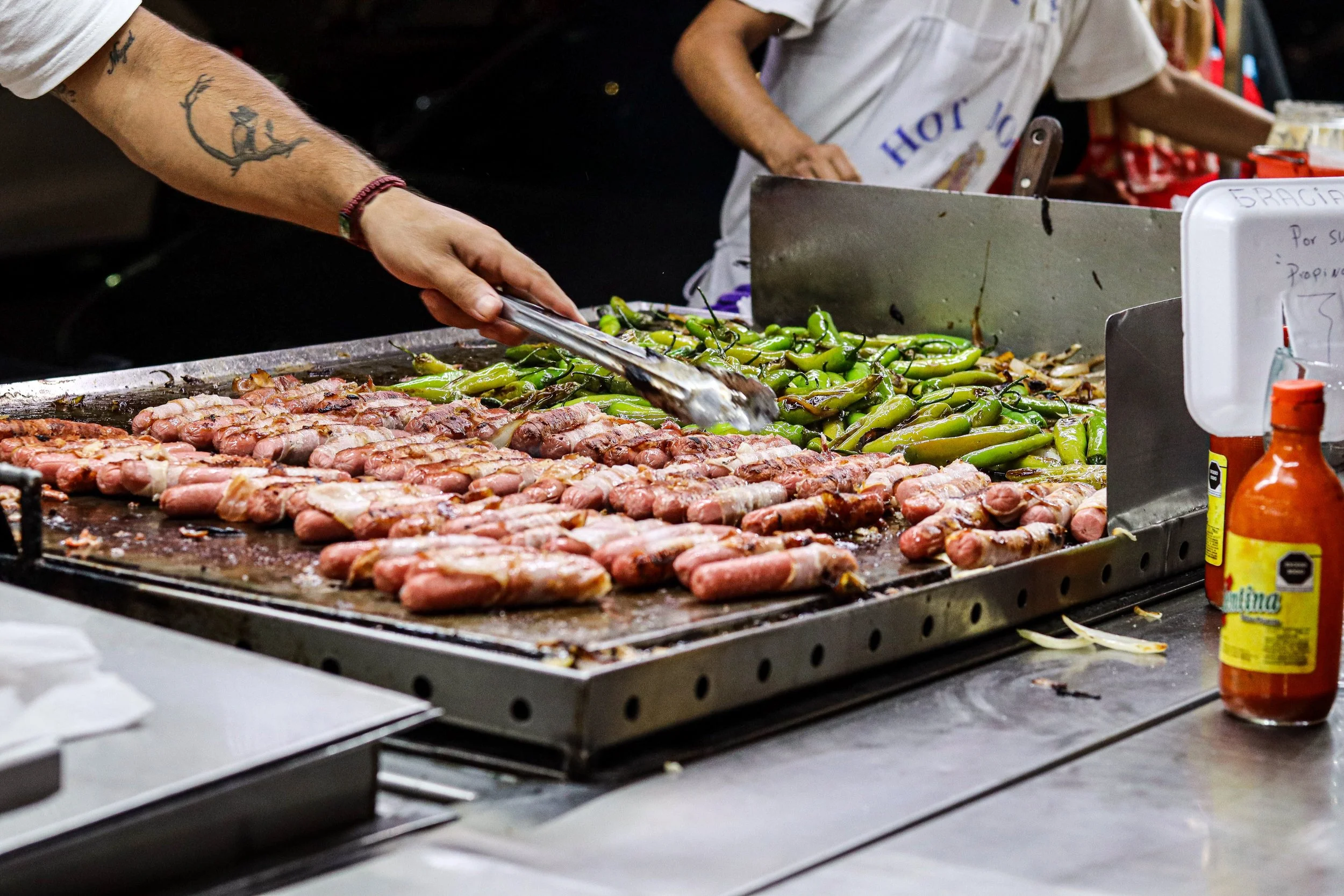 Street food vendor grilling bacon-wrapped jalapenos and green peppers on a large flat griddle.