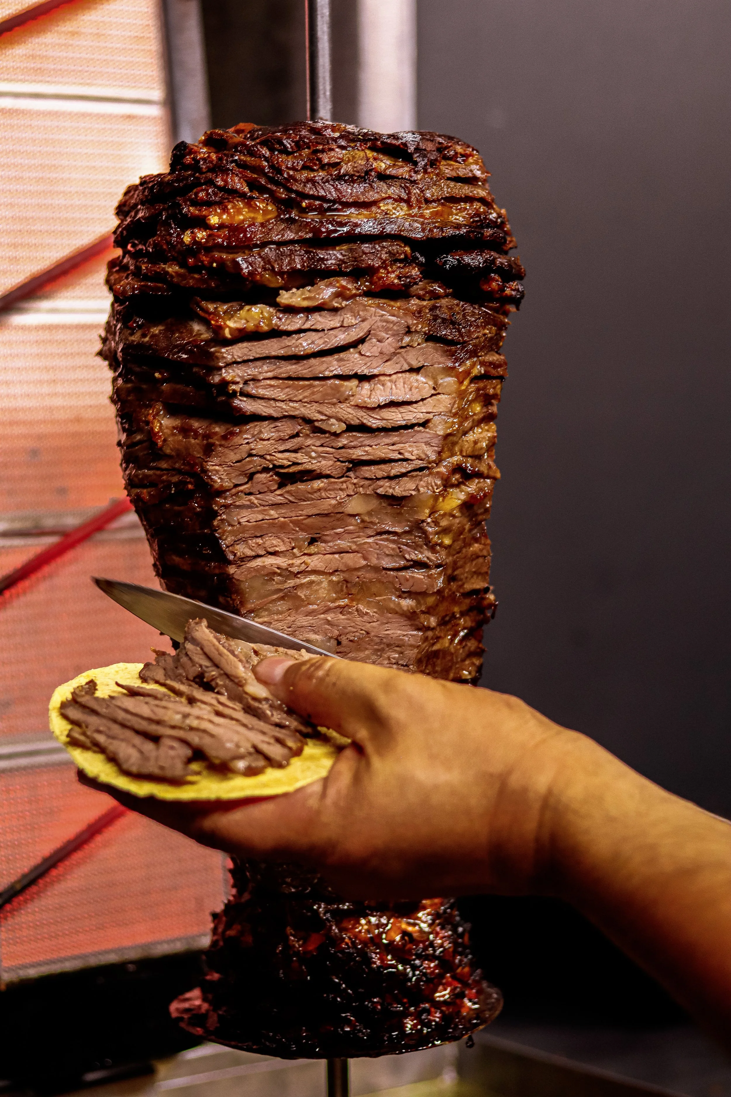A person slices a large, cooked beef brisket. The brisket is mounted on a vertical rotisserie, with a hand holding a knife and a slice of meat on a bun.