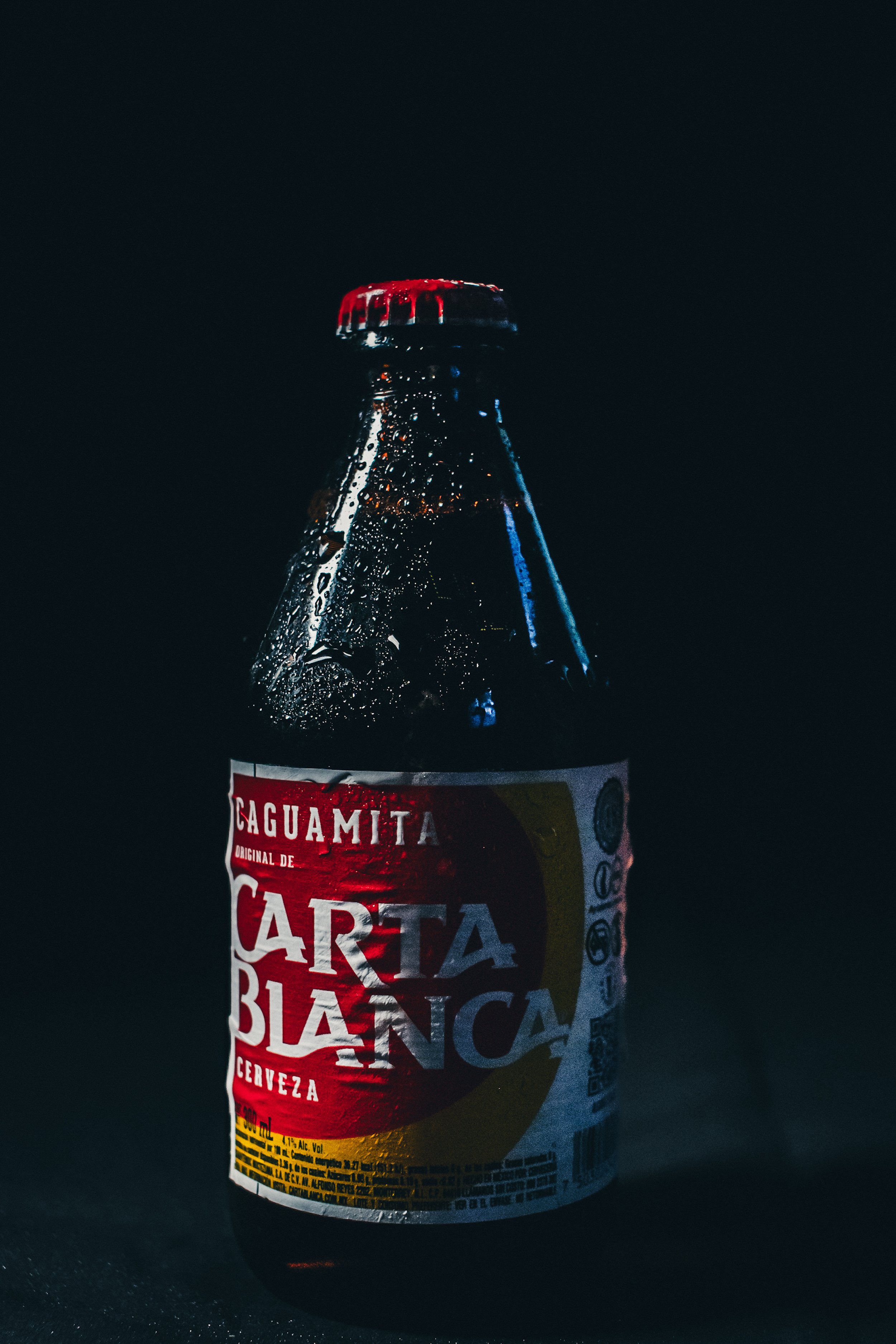 A dark glass bottle of Caguamita Carta Blanca beer with condensation, illuminated against a black background.