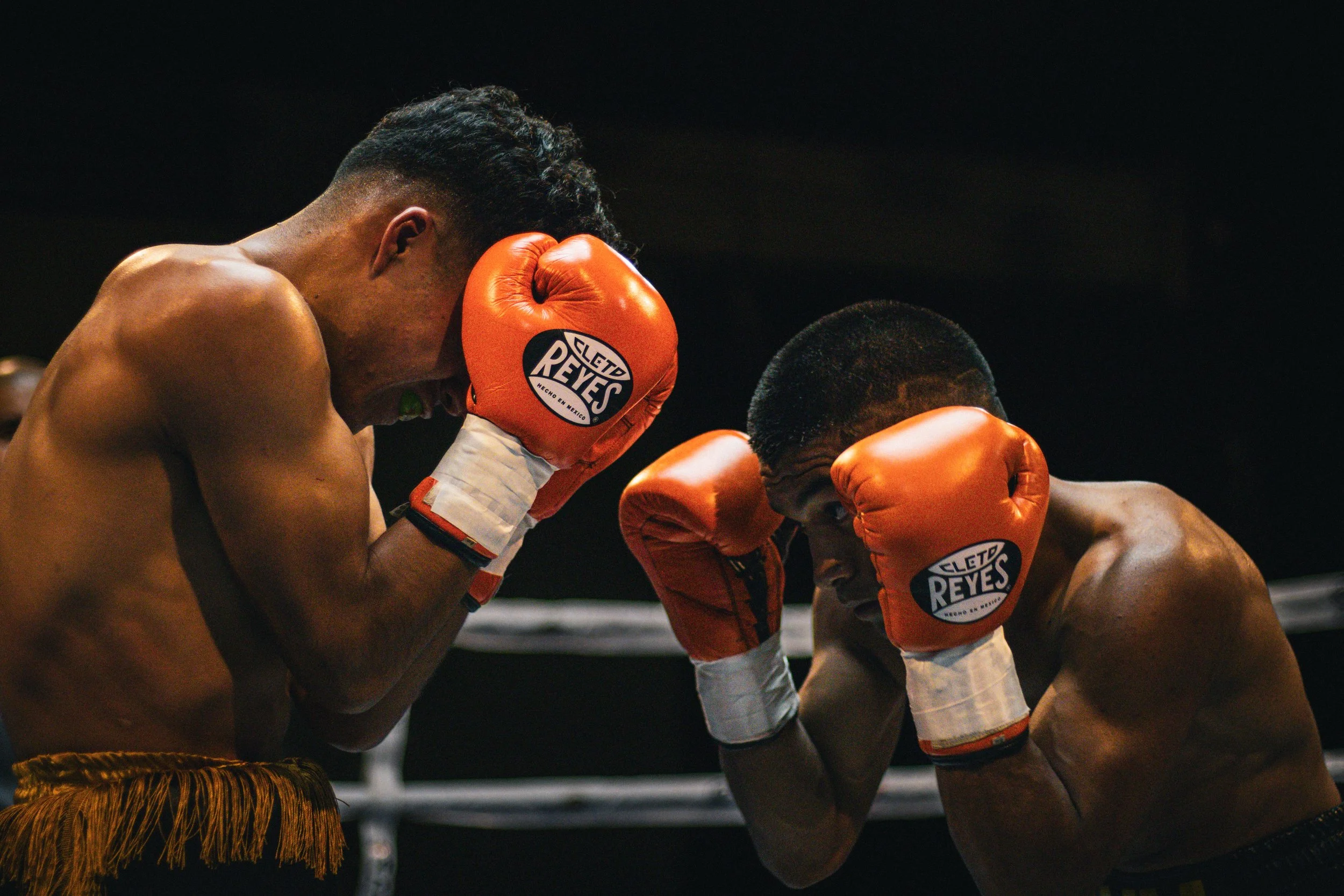 Two shirtless male boxers wearing orange boxing gloves, facing each other in a boxing ring, in a defensive stance during a match.