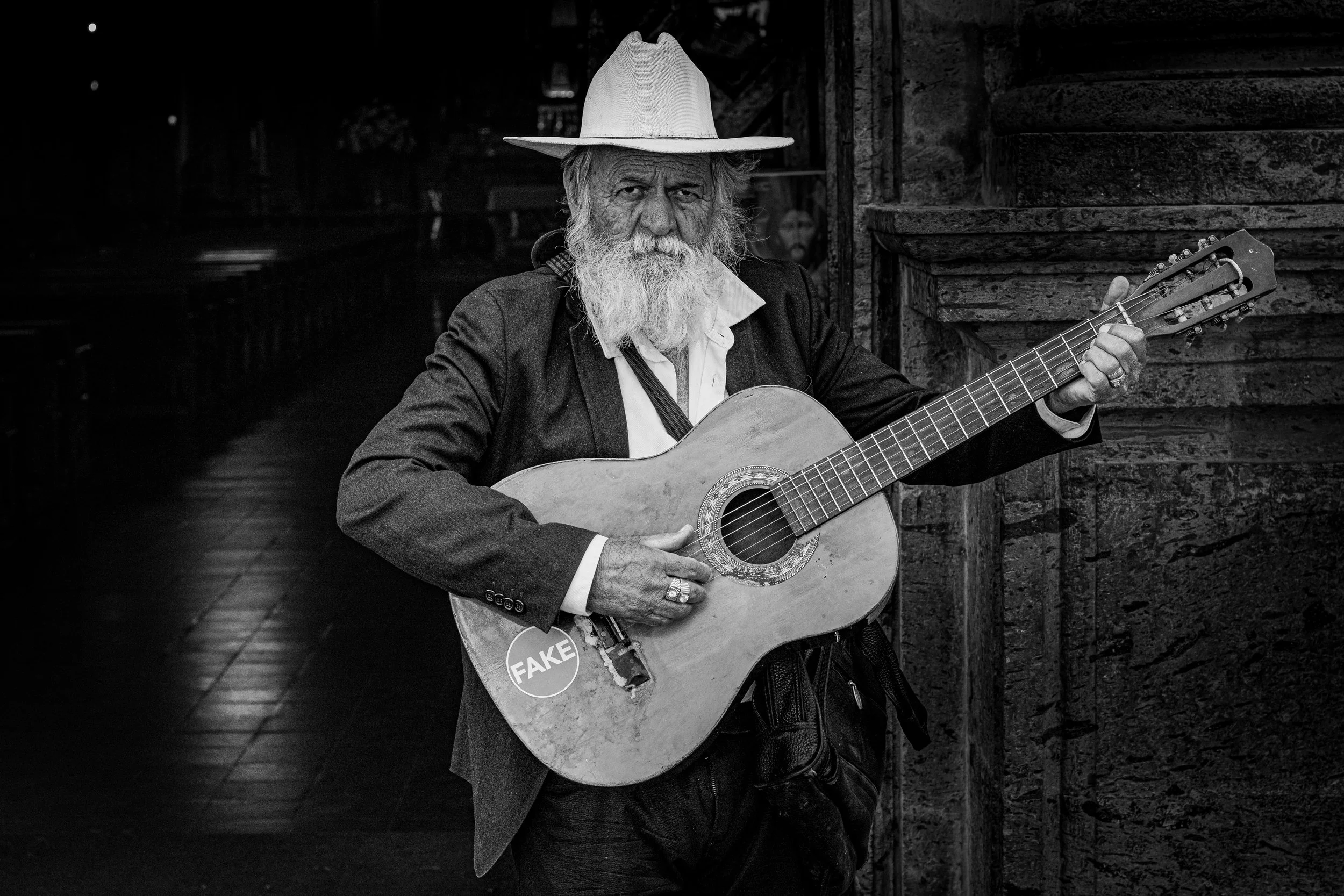 An elderly man with a long white beard and mustache, wearing a wide-brimmed hat and a suit jacket, holding a guitar with a sticker that reads 'FAKE', standing next to a stone building in a dimly lit street.